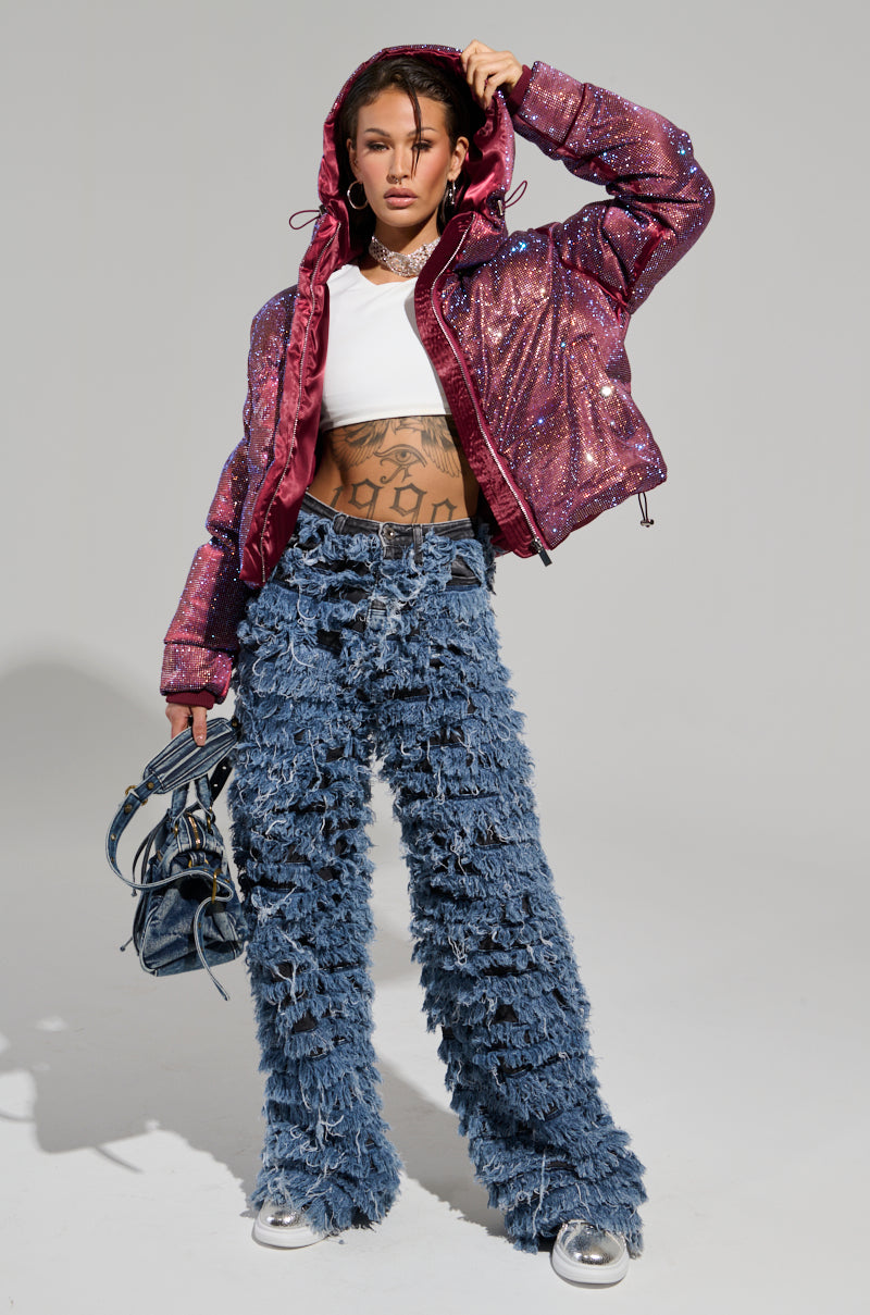A person poses confidently with the hood up in a sparkling red jacket, wearing a white crop top paired with the TOUCH ME DISTRESSED DENIM PANT. They hold a small handbag and wear sneakers, while tattoos are visible on their midriff against a plain, light gray background.
