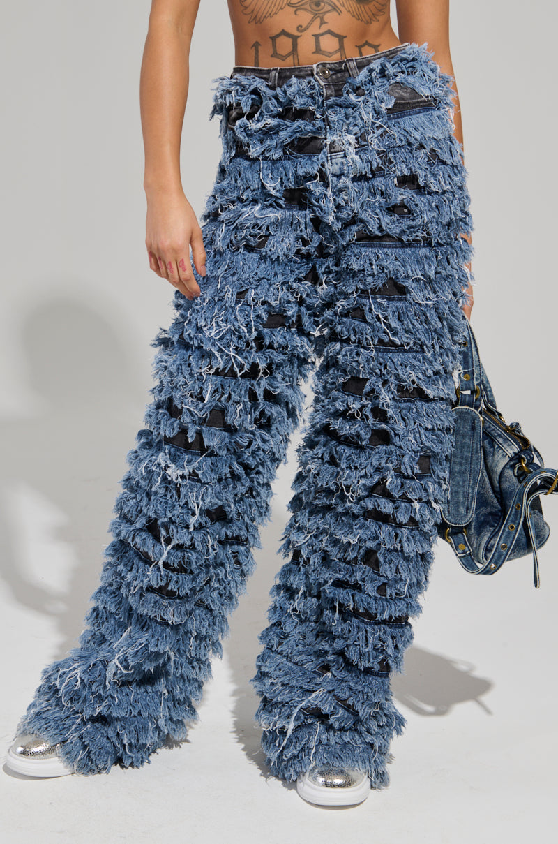 A person is wearing the TOUCH ME DISTRESSED DENIM PANT, featuring heavily fringed, wide-leg jeans, and holding a matching denim bag. Their upper body is partially obscured, revealing only part of the midriff against a plain gray background.