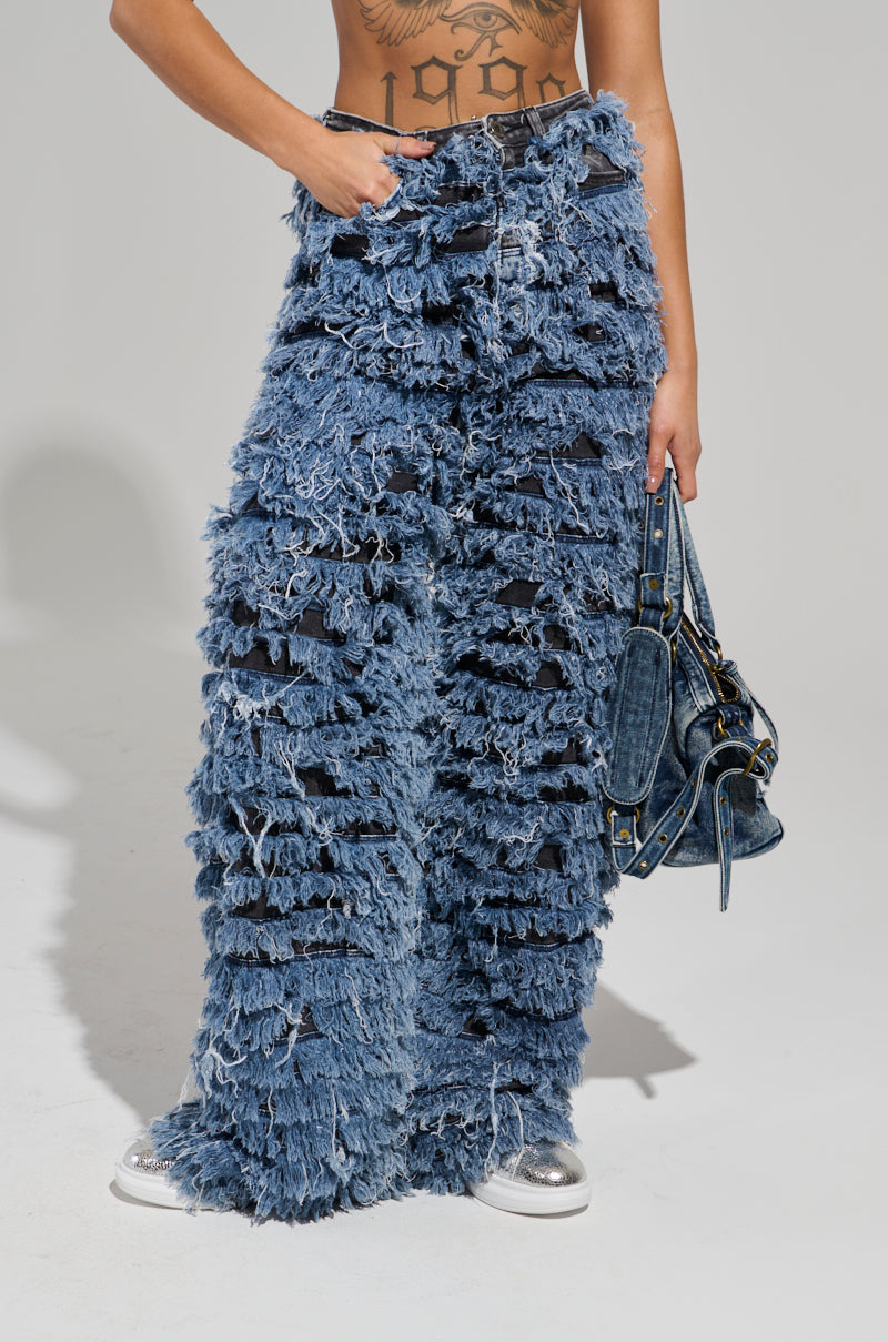 A person stands elegantly in the TOUCH ME DISTRESSED DENIM PANT, paired with a chic blue handbag. Their midriff reveals intricate tattoos, complemented by shiny silver shoes. The plain gray background adds a minimalist touch to the edgy outfit.