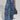 A person stands elegantly in the TOUCH ME DISTRESSED DENIM PANT, paired with a chic blue handbag. Their midriff reveals intricate tattoos, complemented by shiny silver shoes. The plain gray background adds a minimalist touch to the edgy outfit.