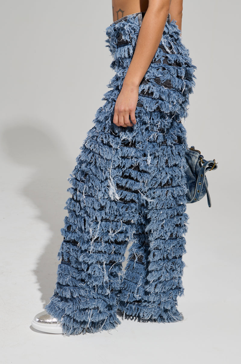 Clad in the TOUCH ME DISTRESSED DENIM PANT, which features a distinctive, heavily fringed wide-leg design and a layered, textured look, an individual walks while holding a small blue handbag. Complementing the outfit with silver shoes, they stand against a plain grey background.