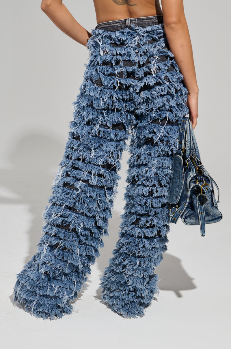 A person wearing the distinctive TOUCH ME DISTRESSED DENIM PANT with high-rise and fringed layers of shredded fabric creates a textured look, complemented by a denim bag against a plain, light grey background.