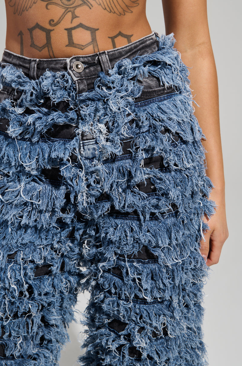 A person is wearing the TOUCH ME DISTRESSED DENIM PANT with frayed, heavily distressed texture and shredded detailing. The high-rise waist of these jeans slightly reveals a tattoo displaying the numbers "1990," against a plain gray backdrop.