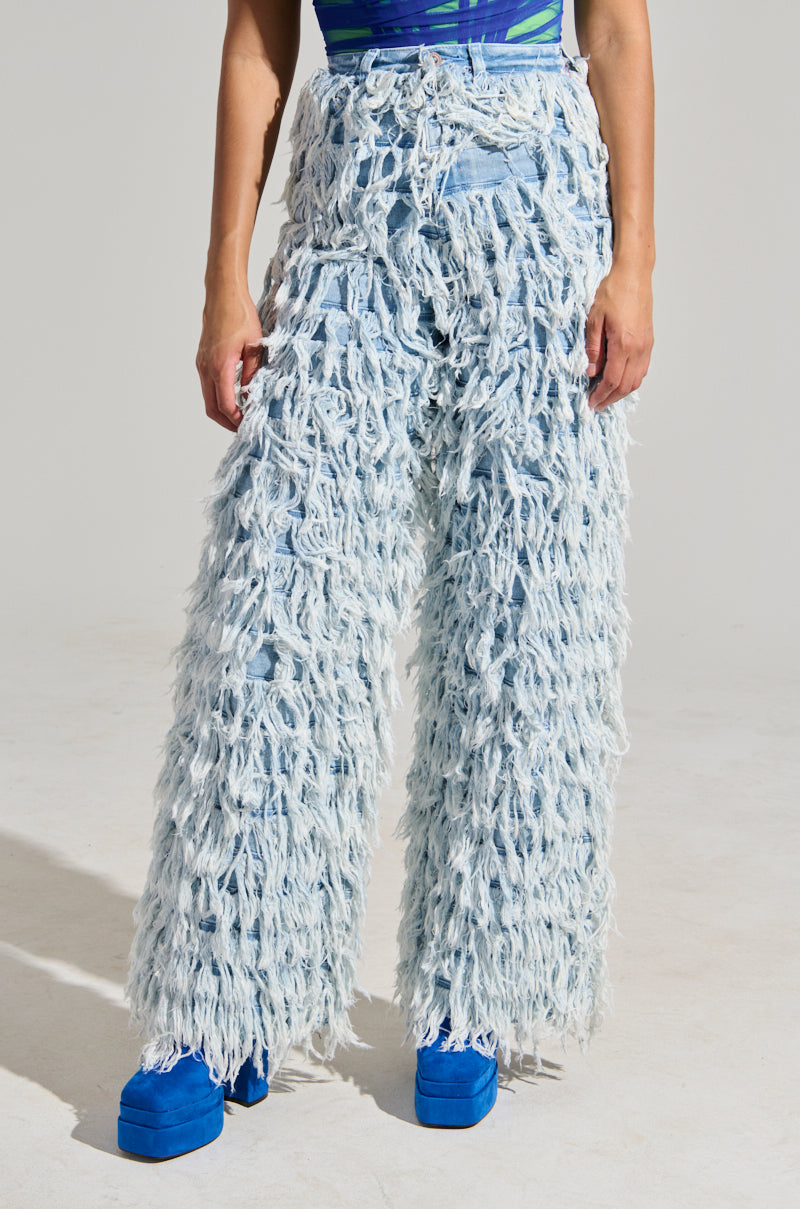 A person in a standout ensemble featuring the BABY ITS YOU DENIM PANT, with its light blue, fringed wide-leg design, paired with blue platform shoes. The top is a patterned tank showcasing blue and green hues against a simple light gray backdrop.
