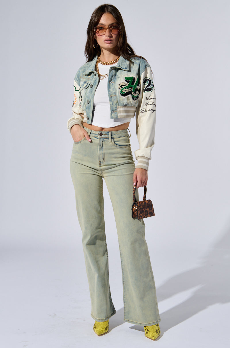 A woman stands confidently against a white background, wearing a cropped, printed jacket over a white top and the Stop Thinking Denim Pant—a pair of high-rise, light-wash jeans with a straight leg silhouette. She accessorizes with sunglasses, a necklace, and a small leopard print handbag while sporting yellow heeled shoes.