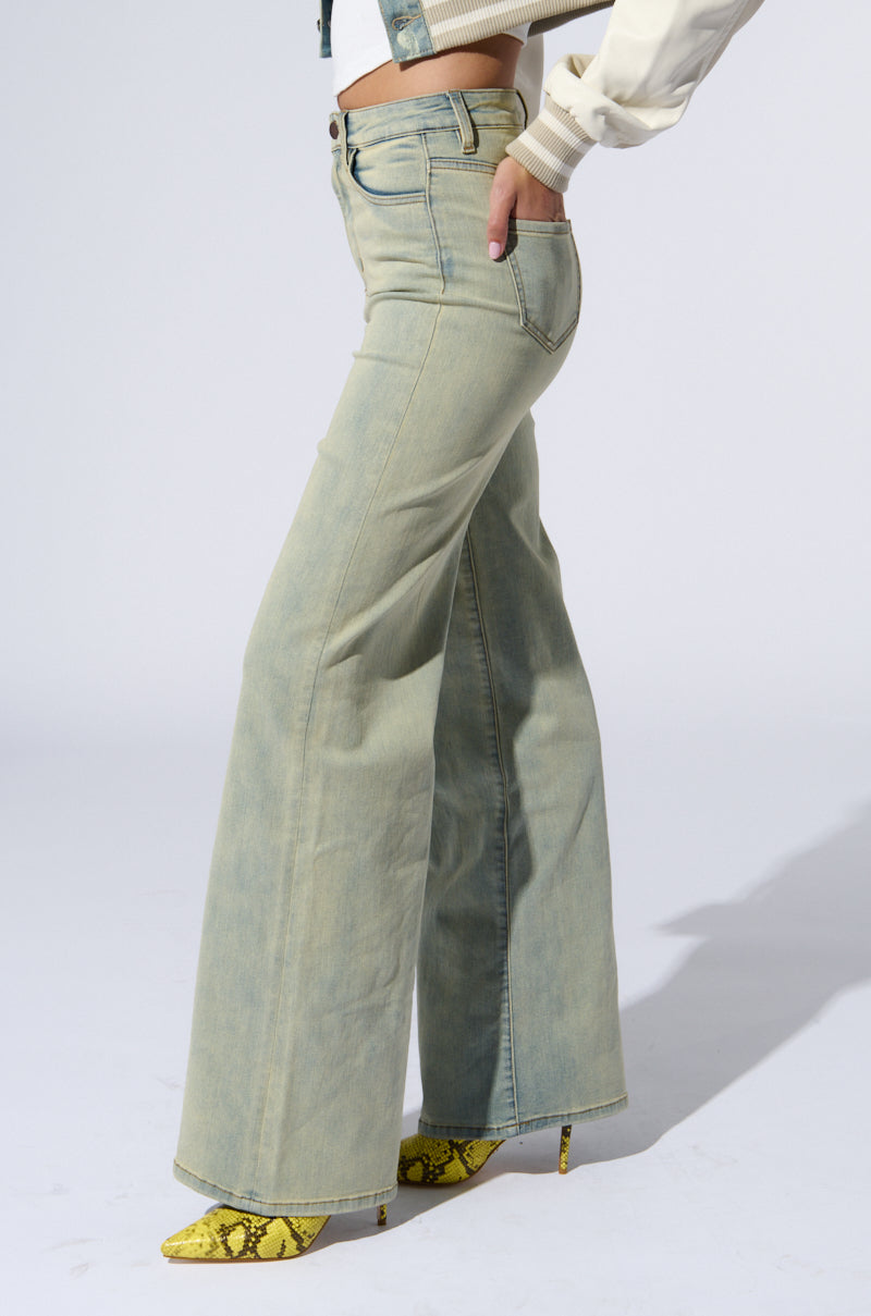 A person wearing light blue STOP THINKING DENIM PANT with a high-rise waist and straight leg silhouette, paired with yellow snakeprint high heels, stands in profile against a plain white background. The denim pants feature classic five-pocket styling and a faded finish, with the person's hand resting casually in their back pocket.