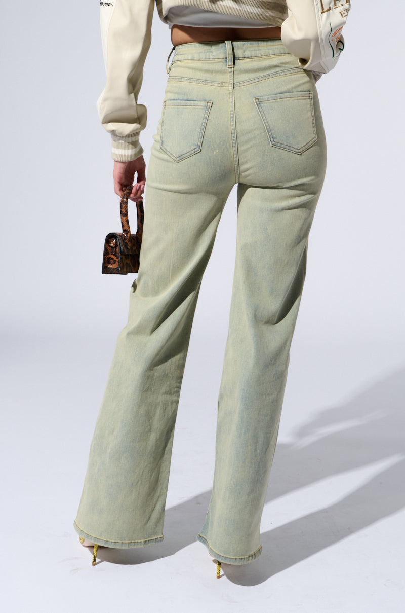 A person is shown from the back, wearing light green STOP THINKING DENIM PANT with a straight-leg silhouette and a beige sweater. They are holding a small, brown, patterned handbag in their left hand. The setting appears to be a studio with a plain, light gray background.