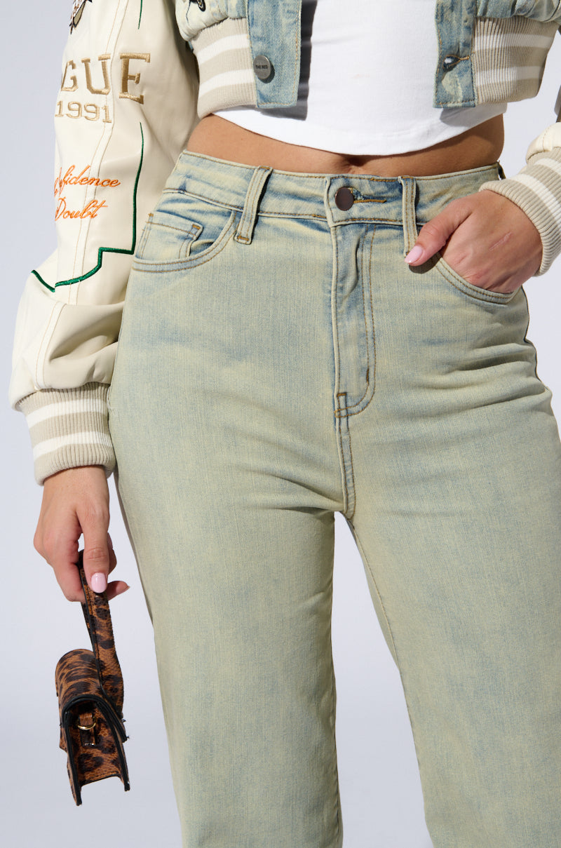 A person dressed in a white crop top and STOP THINKING DENIM PANT holds a small brown patterned bag in one hand. They are also wearing a cream-colored jacket with green text and designs on the sleeve and a denim vest.