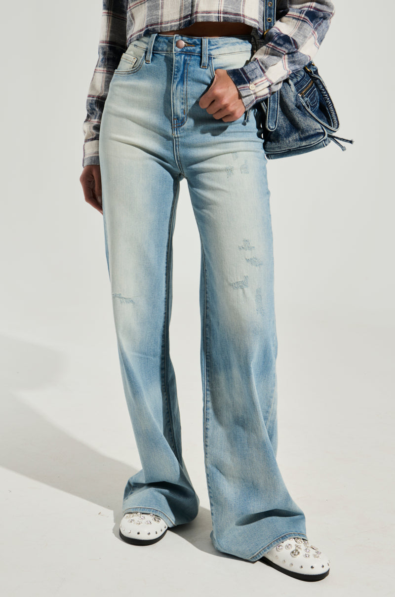 A person wearing "DONT WISH FOR TOO MUCH DENIM PANT" in light blue high-rise denim wide-leg jeans with distressed detailing, a plaid long-sleeve shirt, and white studded shoes. They are holding a small denim handbag. The background is plain and light-colored.