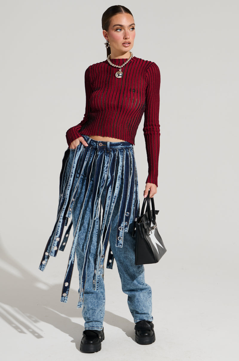 A person wearing a red ribbed long-sleeve top and the NEVER TOO LATE DENIM PANT, featuring blue acid-wash denim with attached strips. They hold a black handbag and wear black shoes. Accessories include a necklace and earrings against a plain, light-colored background.
