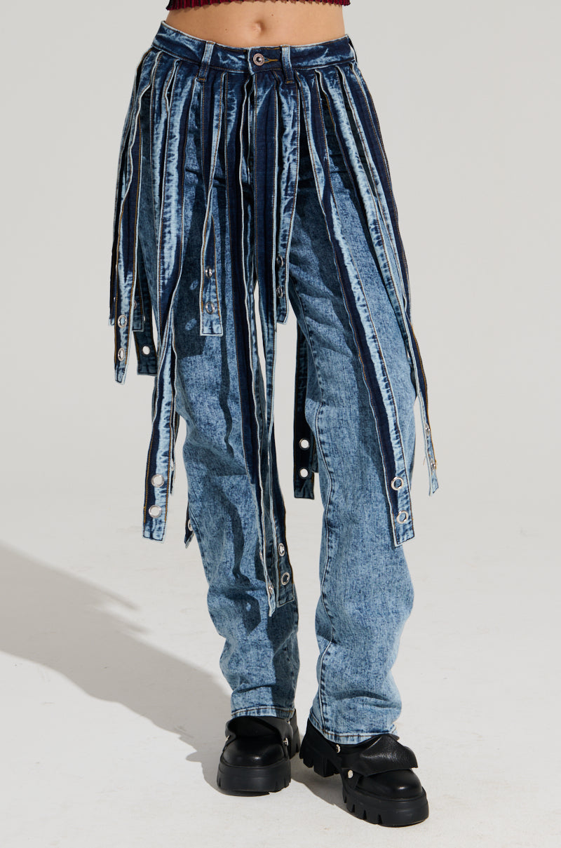 A person wearing the NEVER TOO LATE DENIM PANT featuring high-rise blue denim and multiple attached denim strips, paired with chunky black shoes, stands against a plain background.