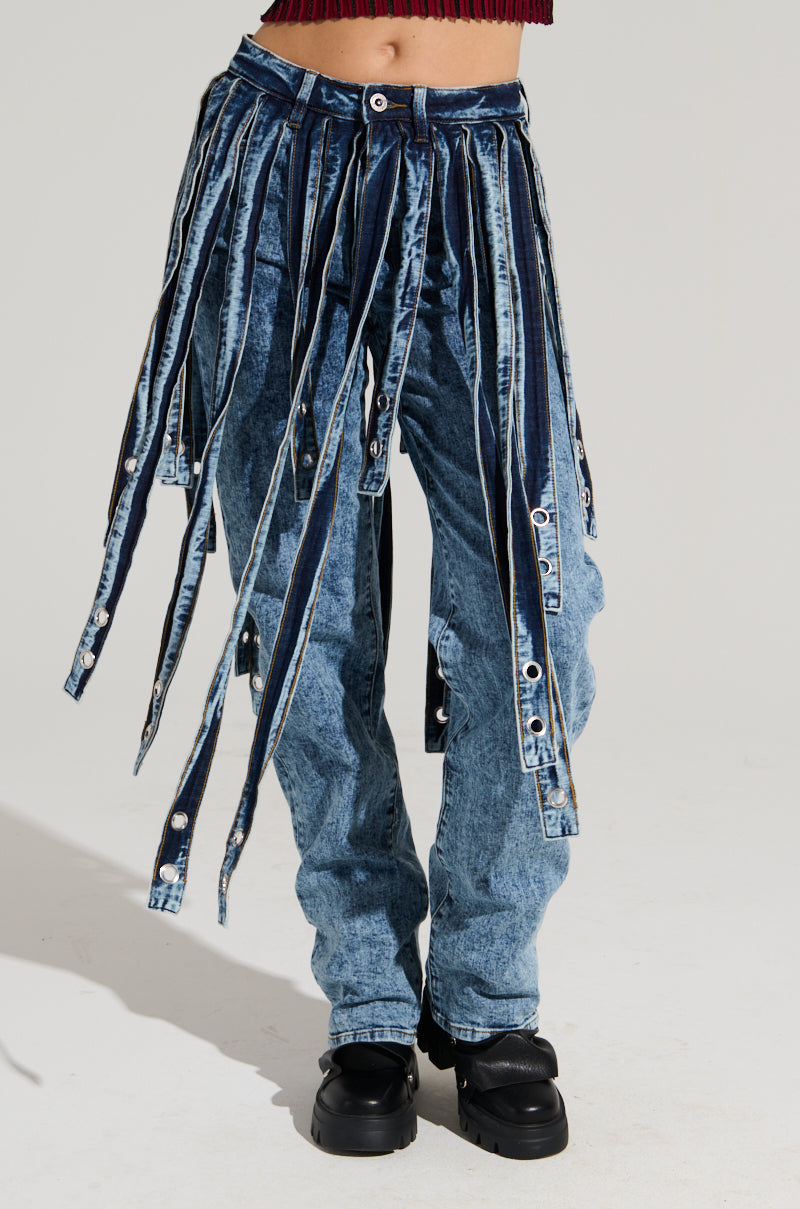A person wears the NEVER TOO LATE DENIM PANT in blue, characterized by multiple extended straps hanging down, paired with black shoes, against a plain white background. The high-rise jeans boast a unique and edgy design, creating a striking visual effect.