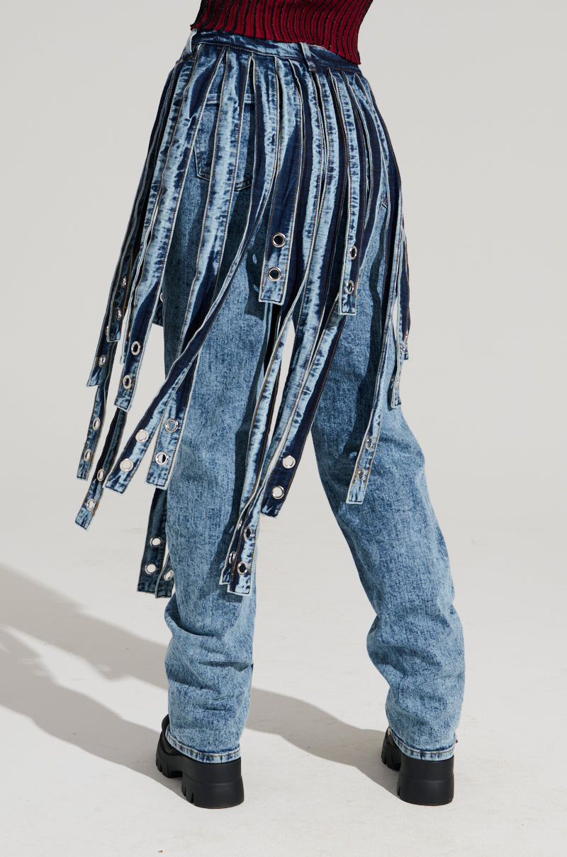 Dressed in the NEVER TOO LATE DENIM PANT, featuring light blue high-rise acid-wash jeans with denim strips cascading from the waist, a person completes their ensemble with black boots and a dark red and black top, all set against a plain light gray background.
