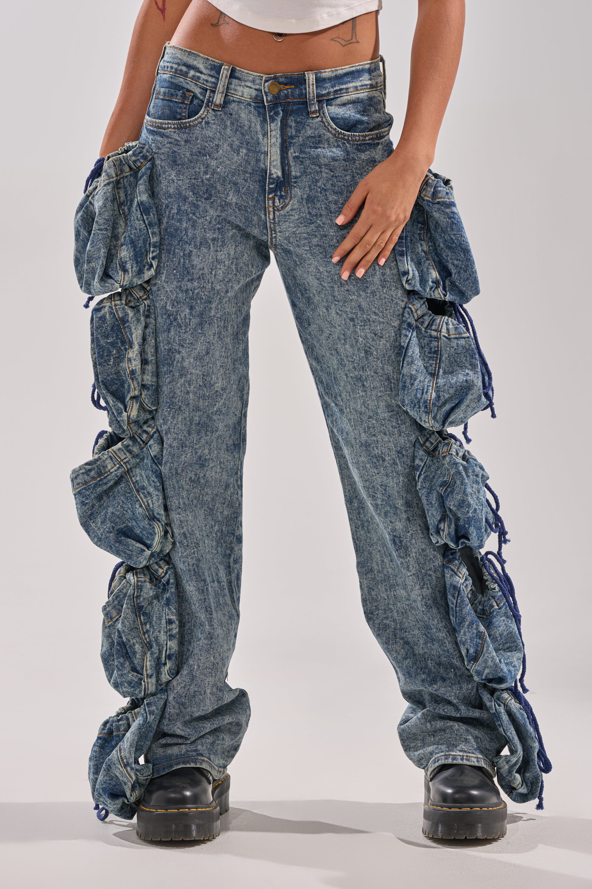 Wearing POCKETS FULL OF SUNSHINE DENIM PANT—high-rise stone-wash jeans with distinctive gathered, ruched side panels—paired with black chunky shoes, lower half shown against a plain light background. 0