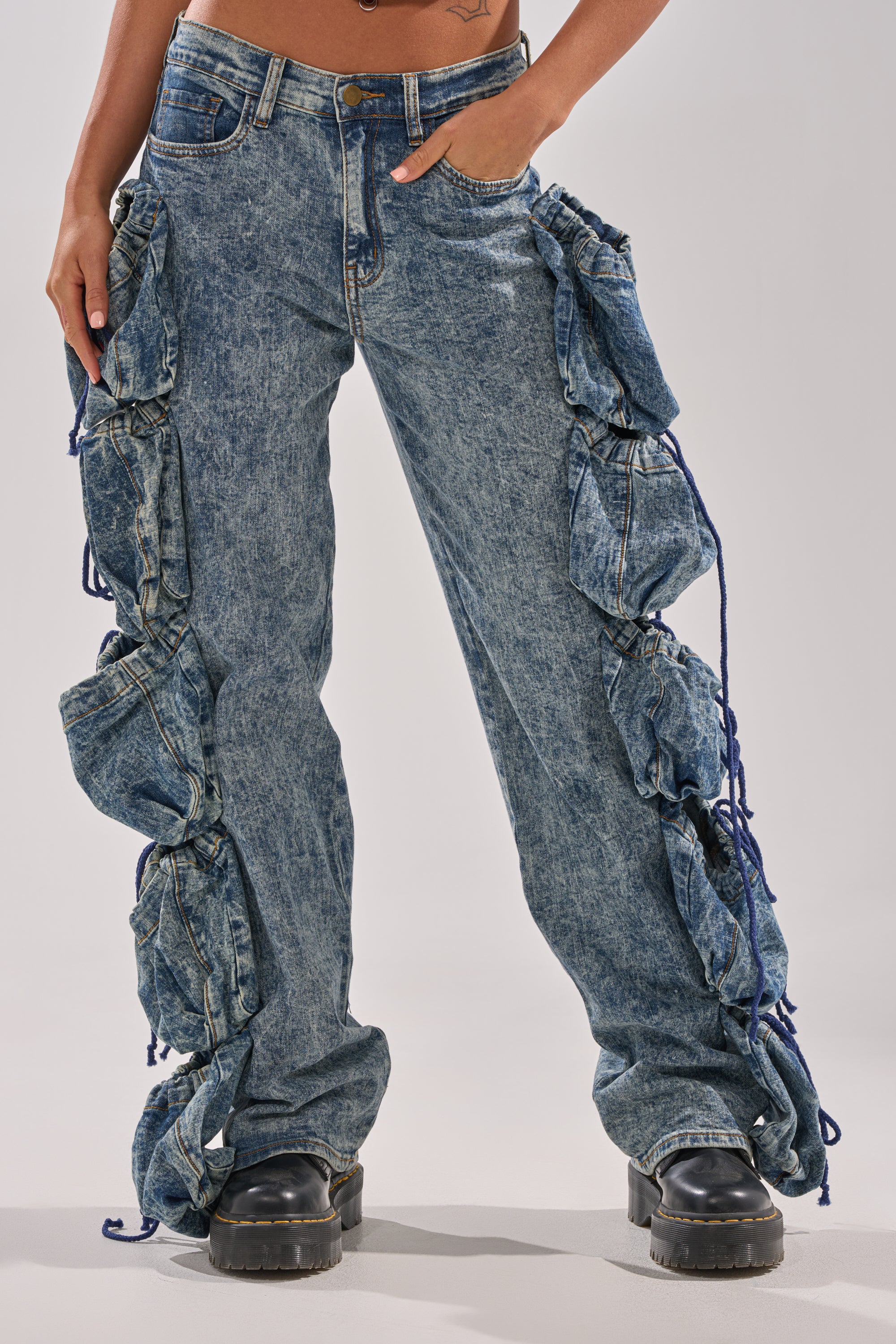 A person in POCKETS FULL OF SUNSHINE DENIM PANT—high-rise acid-wash jeans with large ruched side pockets—pairs them with black platform shoes, standing against a plain light background. 1