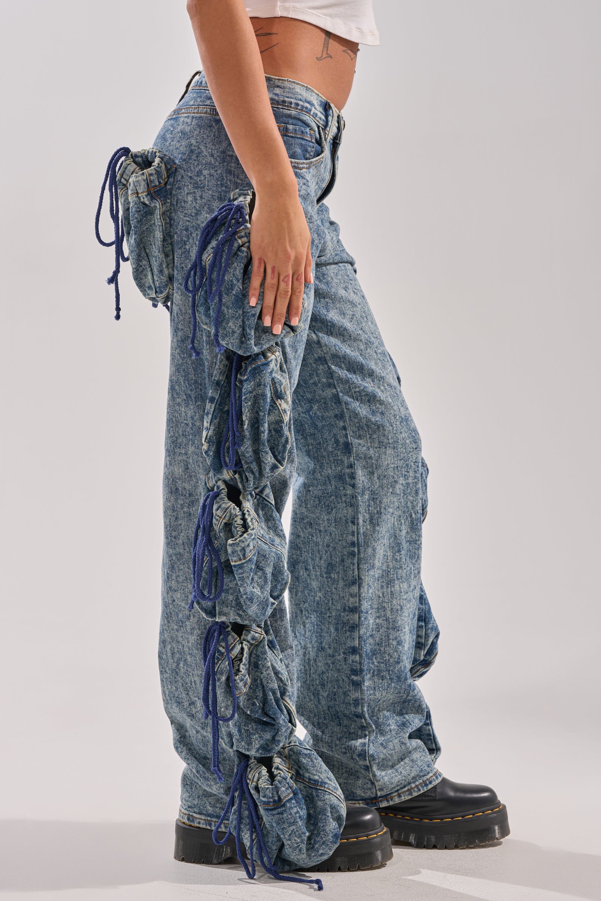 A person wears the POCKETS FULL OF SUNSHINE DENIM PANT—high-rise acid-wash jeans with gathered pockets and blue side drawstrings—paired with black platforms and a white crop top. The photo is side-view against a plain background. 3