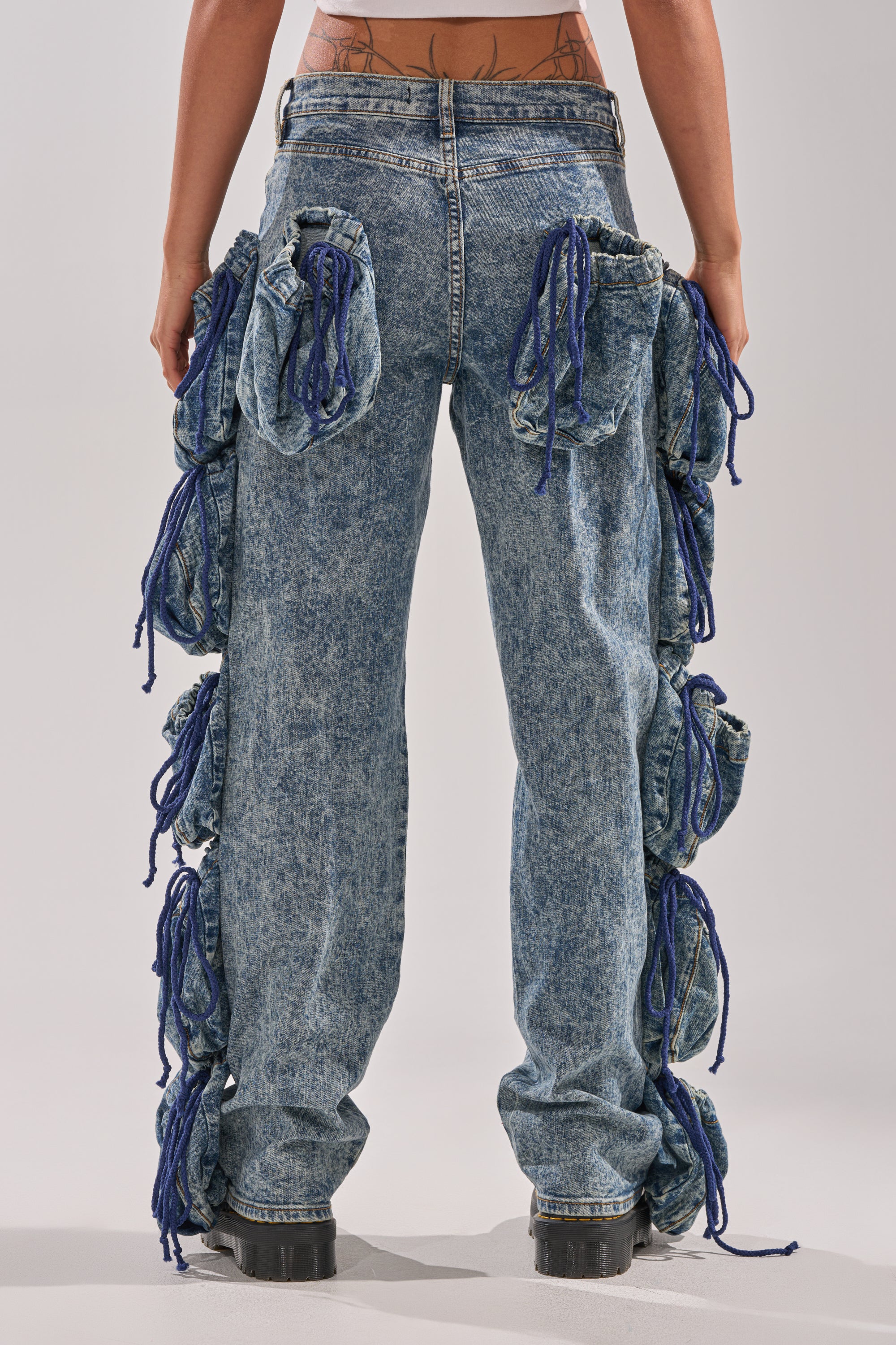 Wearing the POCKETS FULL OF SUNSHINE DENIM PANT—high-rise, acid-wash jeans with large gathered pockets and blue drawstrings—paired with black chunky shoes. Only the lower back and legs are visible. 4