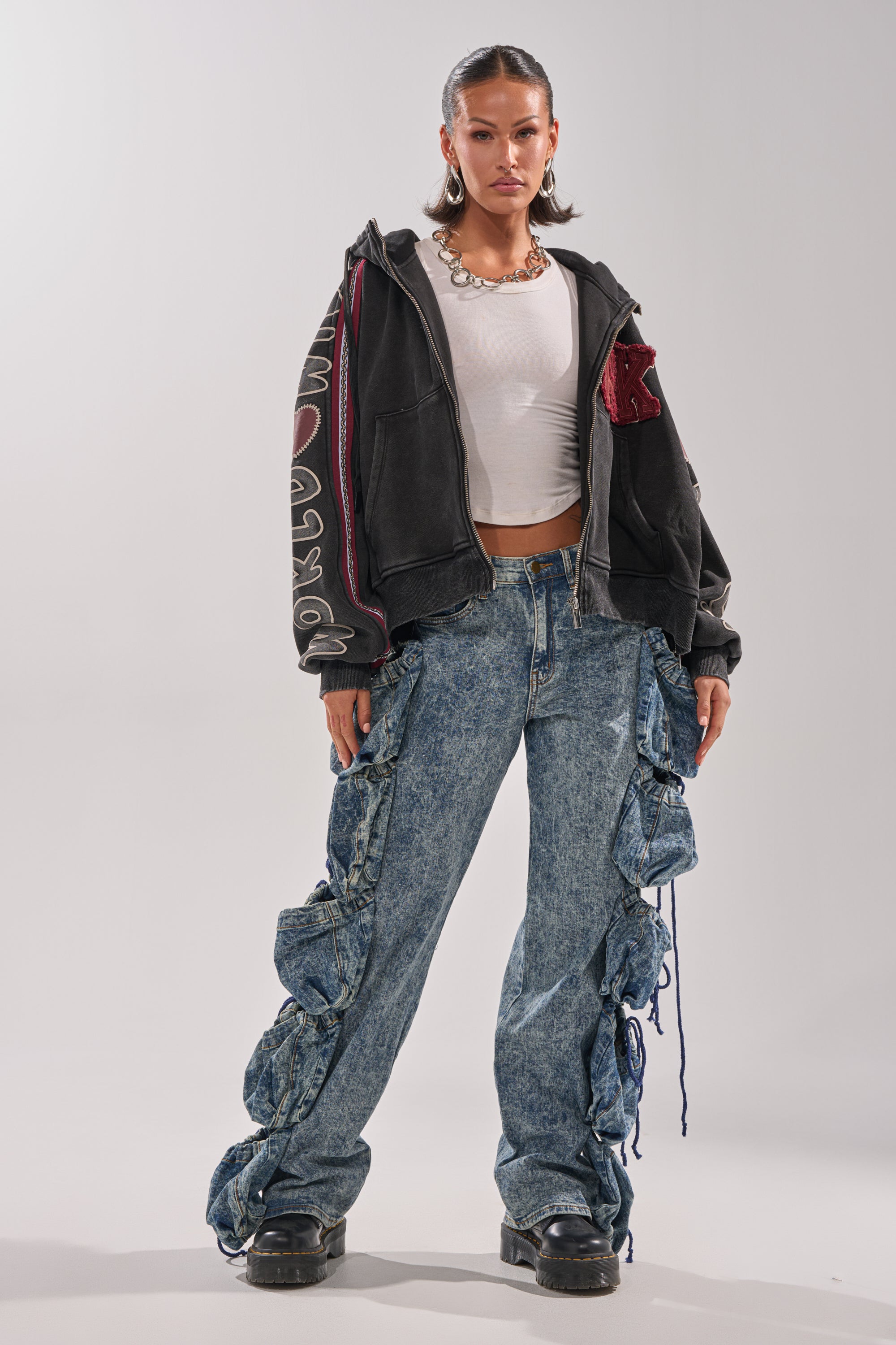A woman faces forward in a black hoodie, white crop top, chunky necklace, hoop earrings, black platform shoes, and blue high-rise "POCKETS FULL OF SUNSHINE DENIM PANT" with ruched, tied side details. 5