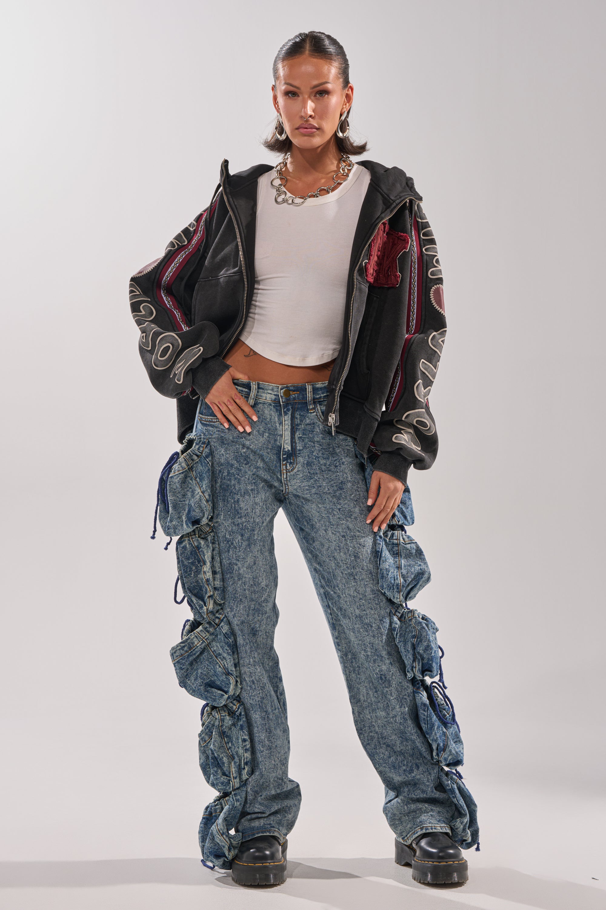 A woman faces forward wearing a black zip-up hoodie with bold white sleeve text, a white crop top, POCKETS FULL OF SUNSHINE DENIM PANT featuring high-rise acid-wash and large ruffled sides, chunky black shoes, and a statement necklace. 6