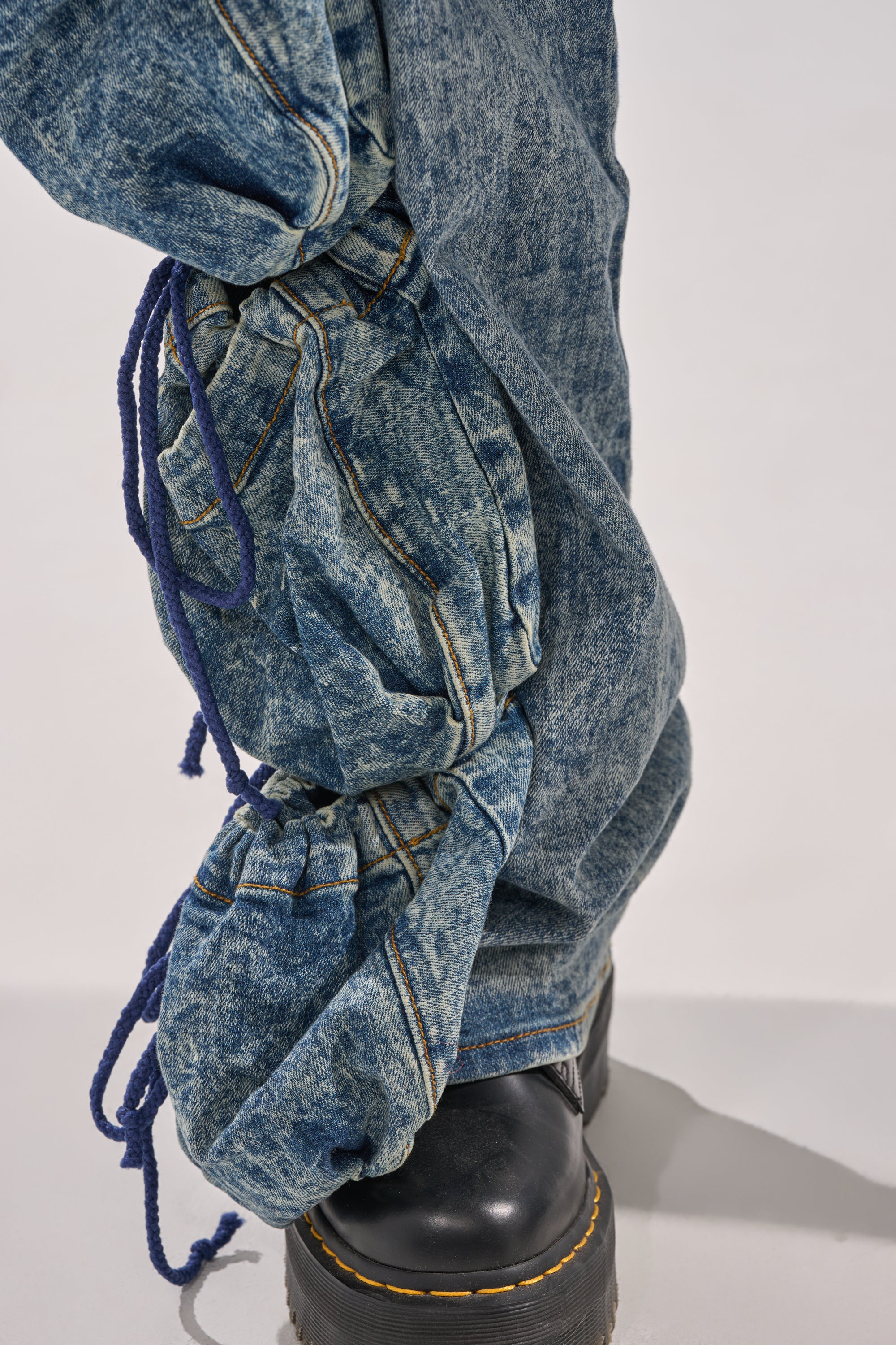 A close-up of a person wearing the POCKETS FULL OF SUNSHINE DENIM PANT—high-rise acid-wash jeans with ruched, blue-tied details and classic five-pocket styling—paired with black leather boots featuring yellow stitching. 8