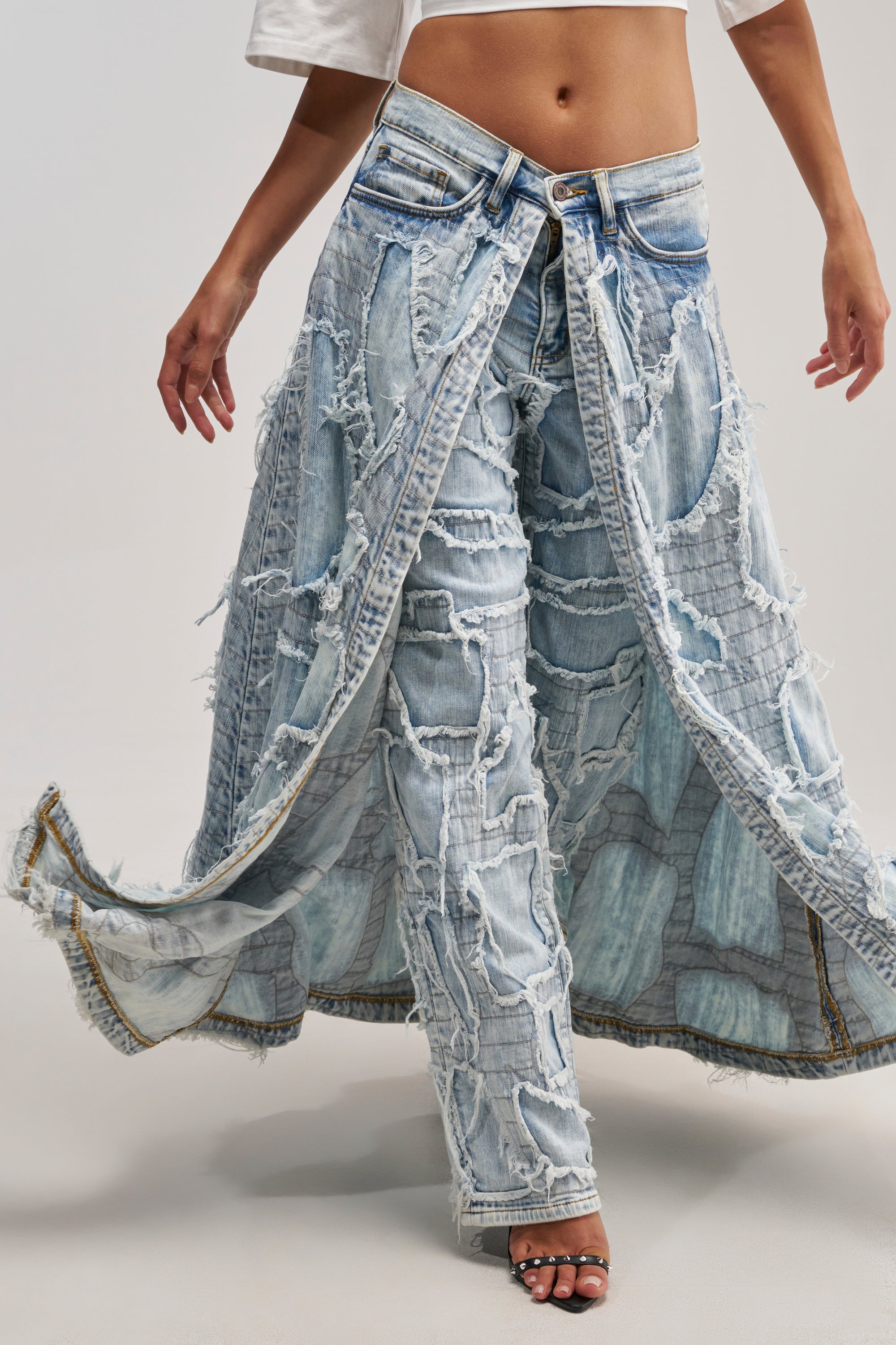 Wearing a white crop top and the CRAZY FOR YOU SUPER DISTRESSED DENIM pants, featuring dramatic frayed panels and layered ripped details for a bold streetwear look, the person stands with arms out and one foot forward.