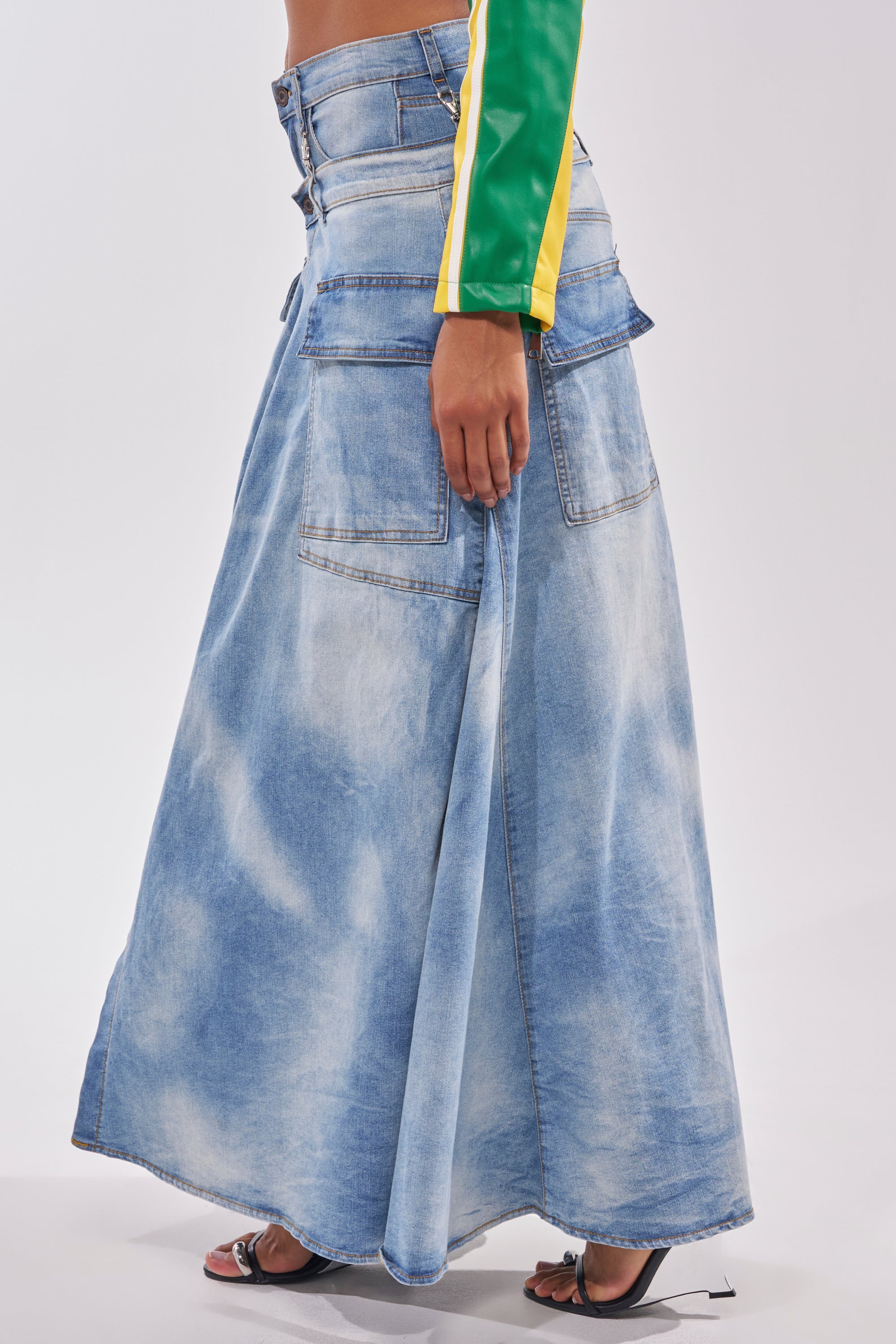 Someone wearing the OVER IT DENIM PANT, a long blue street-style denim maxi skirt with large back pockets, paired with a green and yellow long-sleeve top and black heeled sandals. Side view against a plain background. 4
