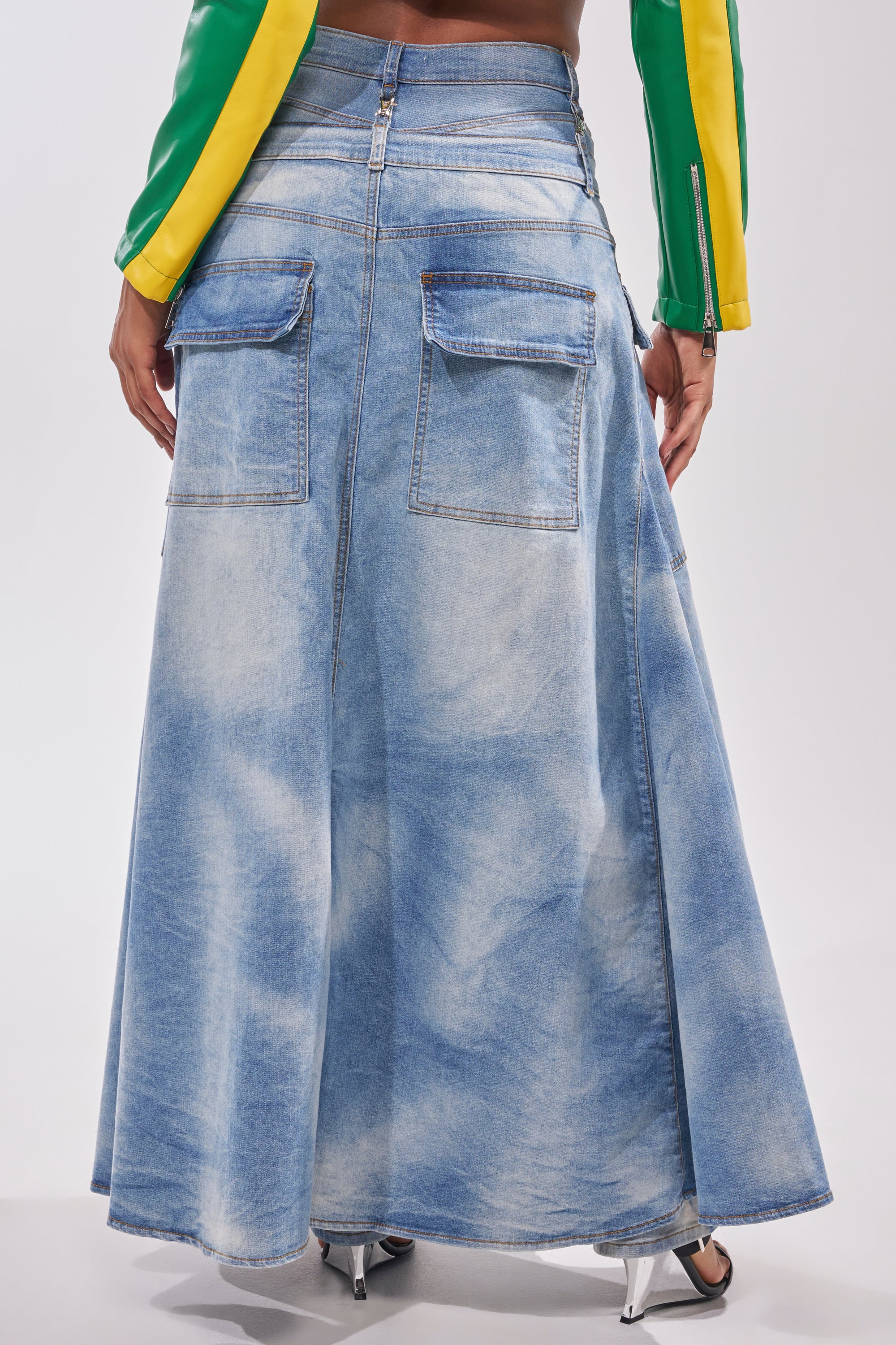 Wearing the OVER IT DENIM PANT, this person exudes deconstructed street style in a long, light blue denim skirt with large back pockets, paired with a green and yellow jacket and silver heels against a white background. 5
