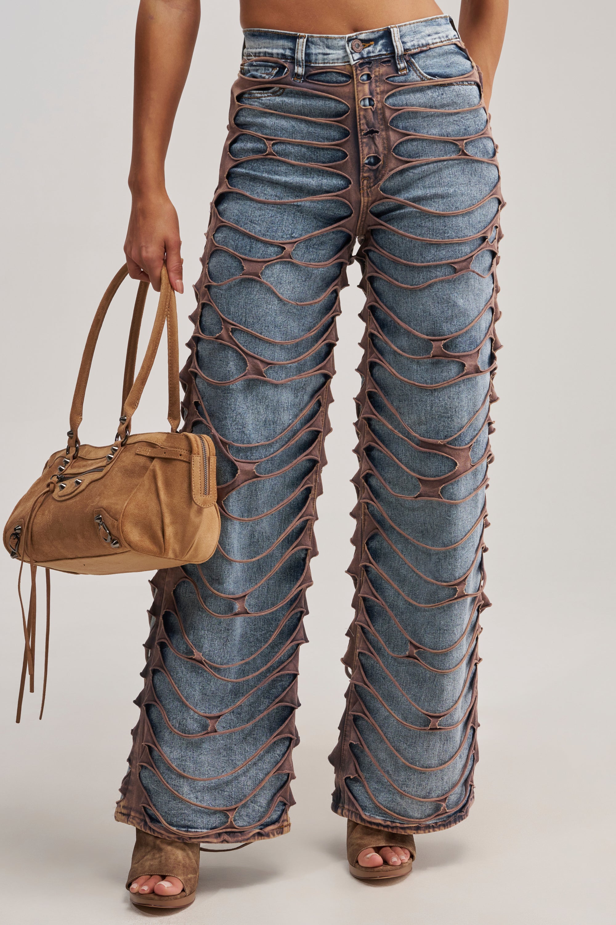 Wearing the TIED TO YOU DENIM PANT—high-waisted, distressed flared-leg jeans with bold cutouts—styled with heels and a tan suede handbag for an edgy-chic look.