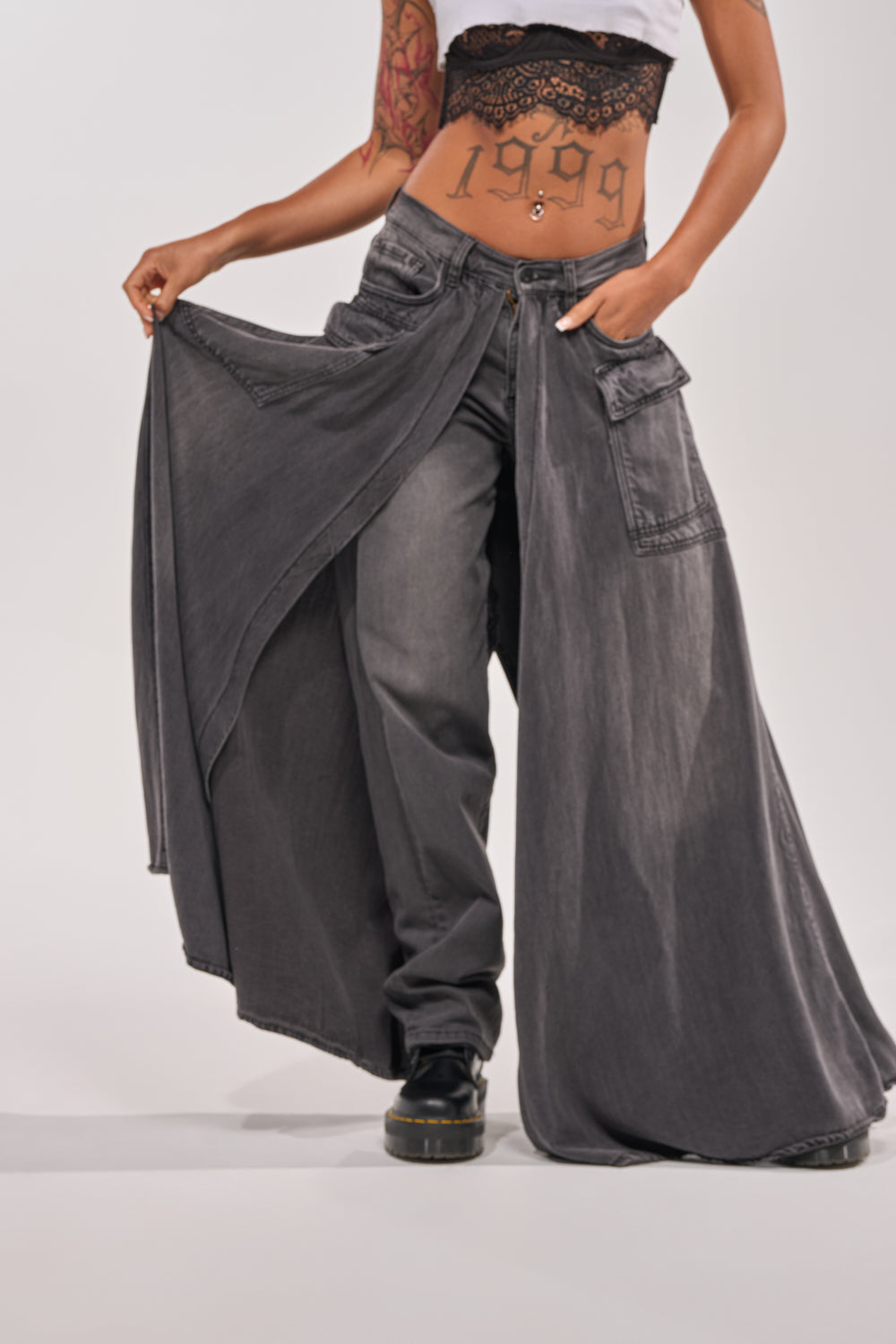 A person wears the MIXED EMOTIONS DENIM PANT, a black lace crop top, and black shoes. Their arms and midriff, showing "1999" and tattoos, stand out against a plain light background. 0