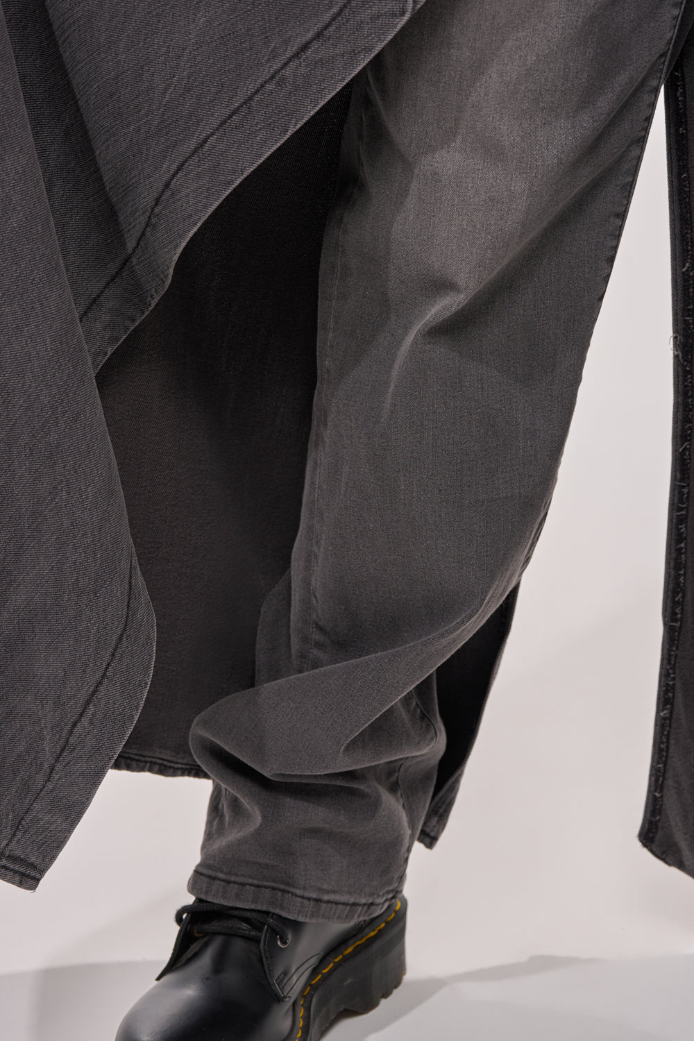 Close-up of a person wearing the MIXED EMOTIONS DENIM PANT with a matching long coat and black leather lace-up boots with yellow stitching, standing on a light surface. 10