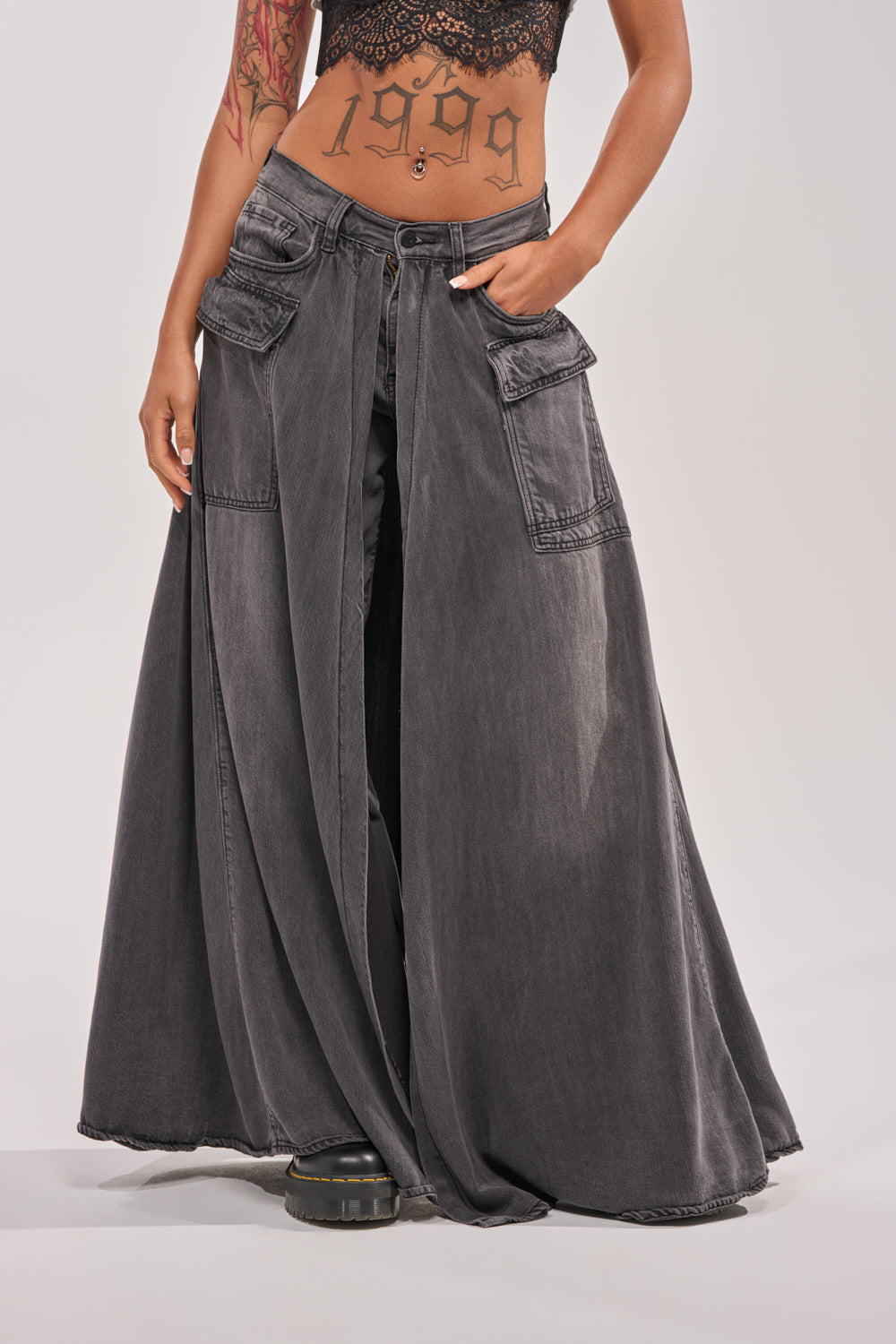 A person wears a black lace crop top and MIXED EMOTIONS DENIM PANT—high-waisted, wide-leg black denim pants with large front pockets and a stylish 2-in-1 skirt overlay. Their midriff tattoos are visible. 1