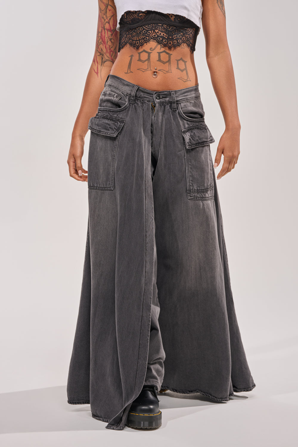 Person wears MIXED EMOTIONS DENIM PANT—wide-leg dark gray cargo jeans with large side pockets—layered with a black denim maxi skirt, a lacy black crop top, and black boots. Arm tattoos and "1999" tattooed on stomach; face not visible. 2