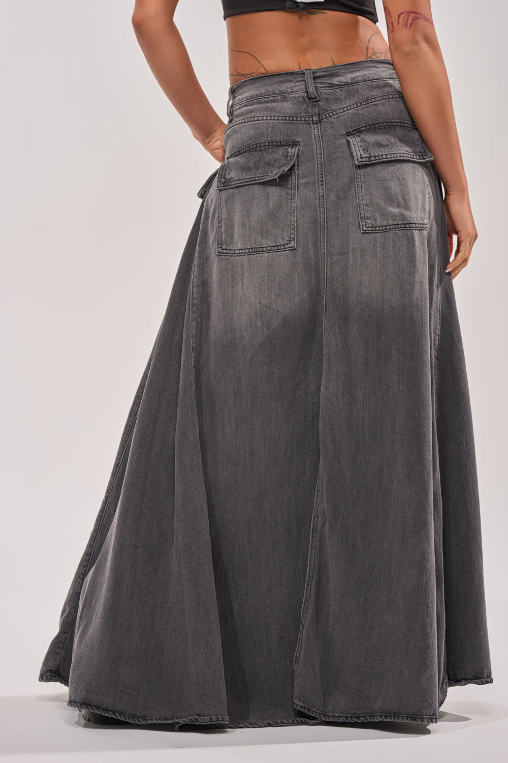 A person wears the MIXED EMOTIONS DENIM PANT, a long flared black denim maxi skirt with large back pockets and raw hem, shown from the back with their hands by their sides. 5