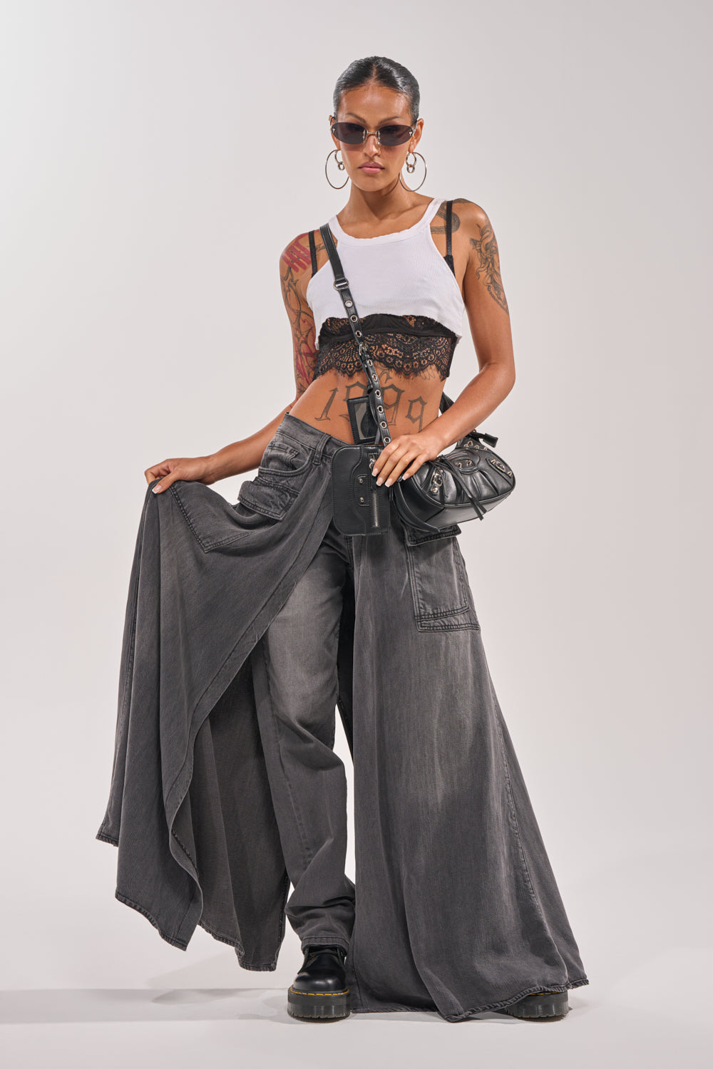 Wearing sunglasses, a white cropped tank over a black lace bralette, MIXED EMOTIONS DENIM PANT, black platform shoes, hoop earrings, and with visible tattoos, this confident model poses stylishly. 6