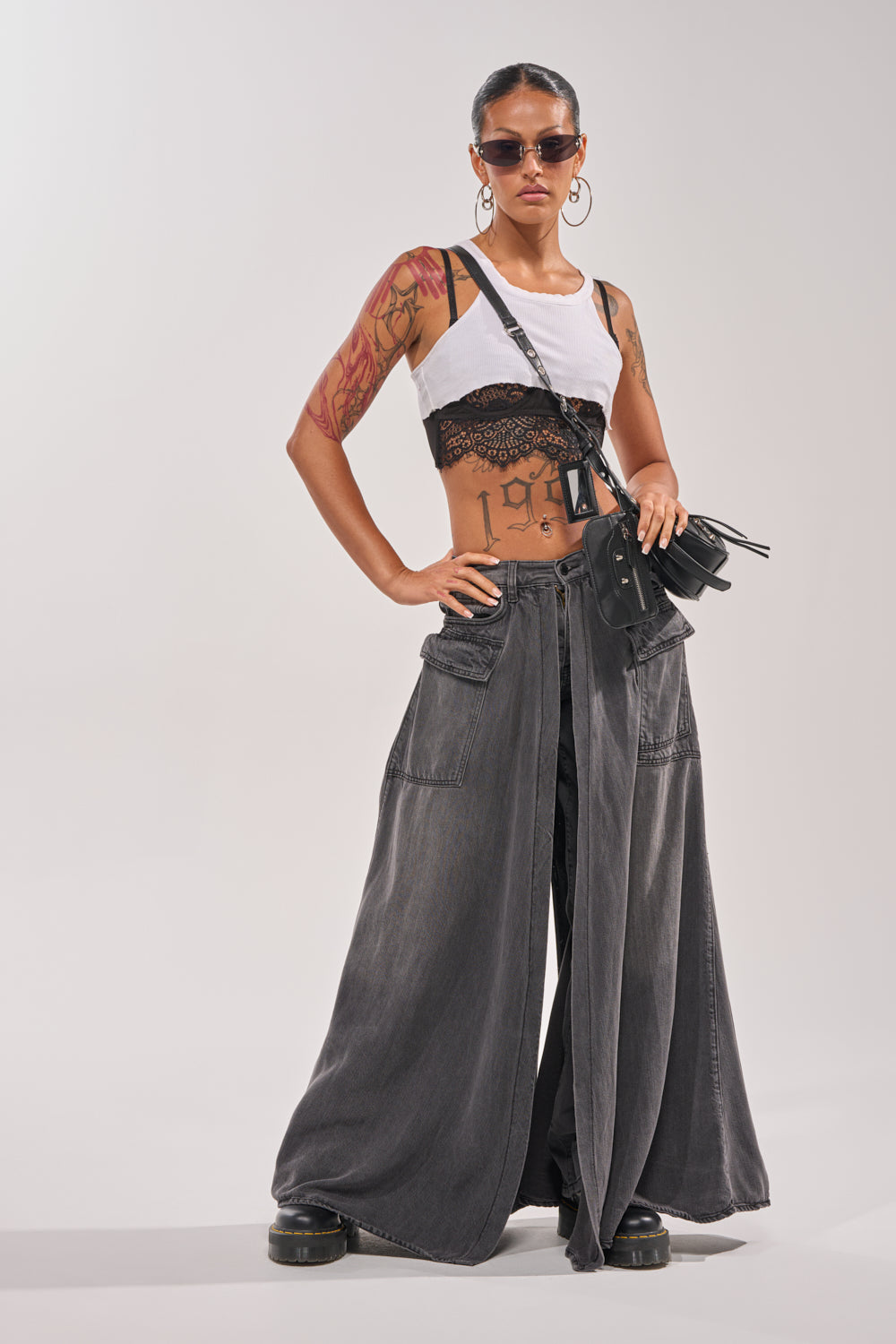 A person with tattoos poses confidently in MIXED EMOTIONS DENIM PANT, a cropped white tank, black lace bralette, chunky black shoes, sunglasses, and a small black bag. 7