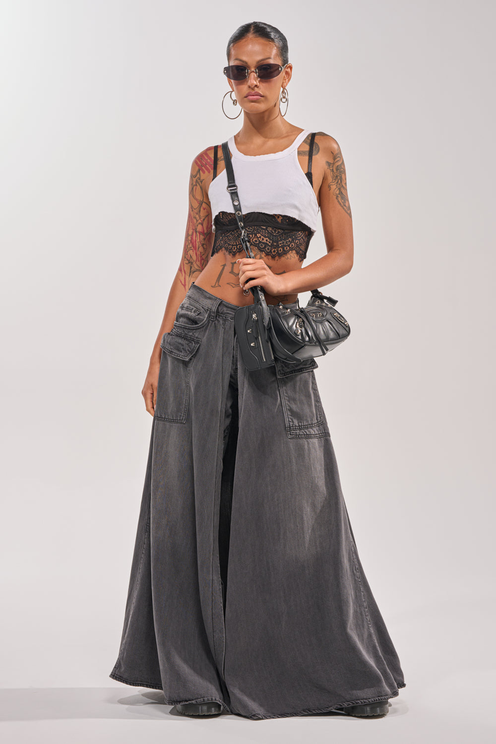 A woman stands confidently against a plain background wearing the MIXED EMOTIONS DENIM PANT, styled with straight-leg pockets, a white crop top over a black lace bralette, sunglasses, large hoop earrings, and a black crossbody handbag. 8