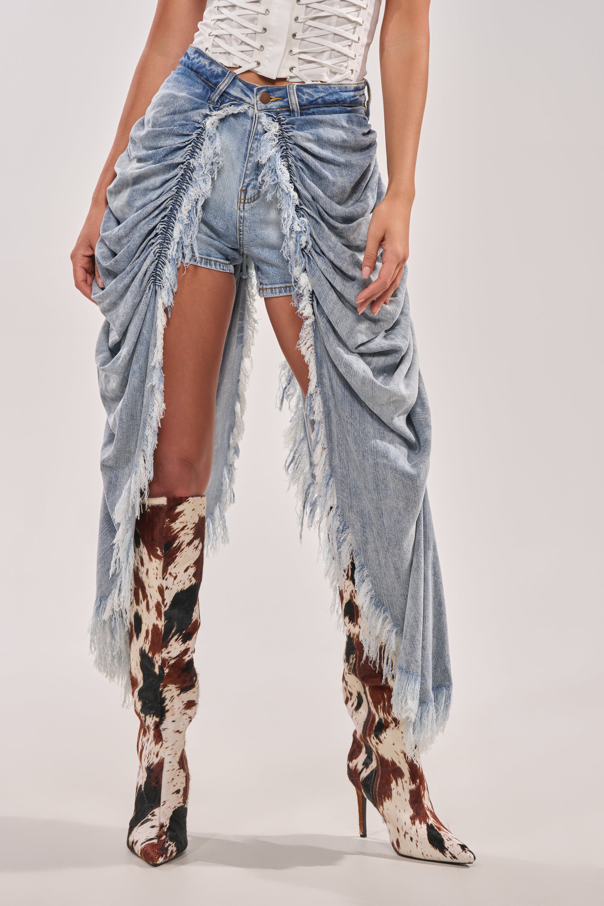 A person wears a white corset top, ALL OVER THE PLACE DENIM SHORT (light-wash, high-rise), a draped, fringed denim skirt, and cow-print heeled boots. The image is cropped at the shoulders. 1
