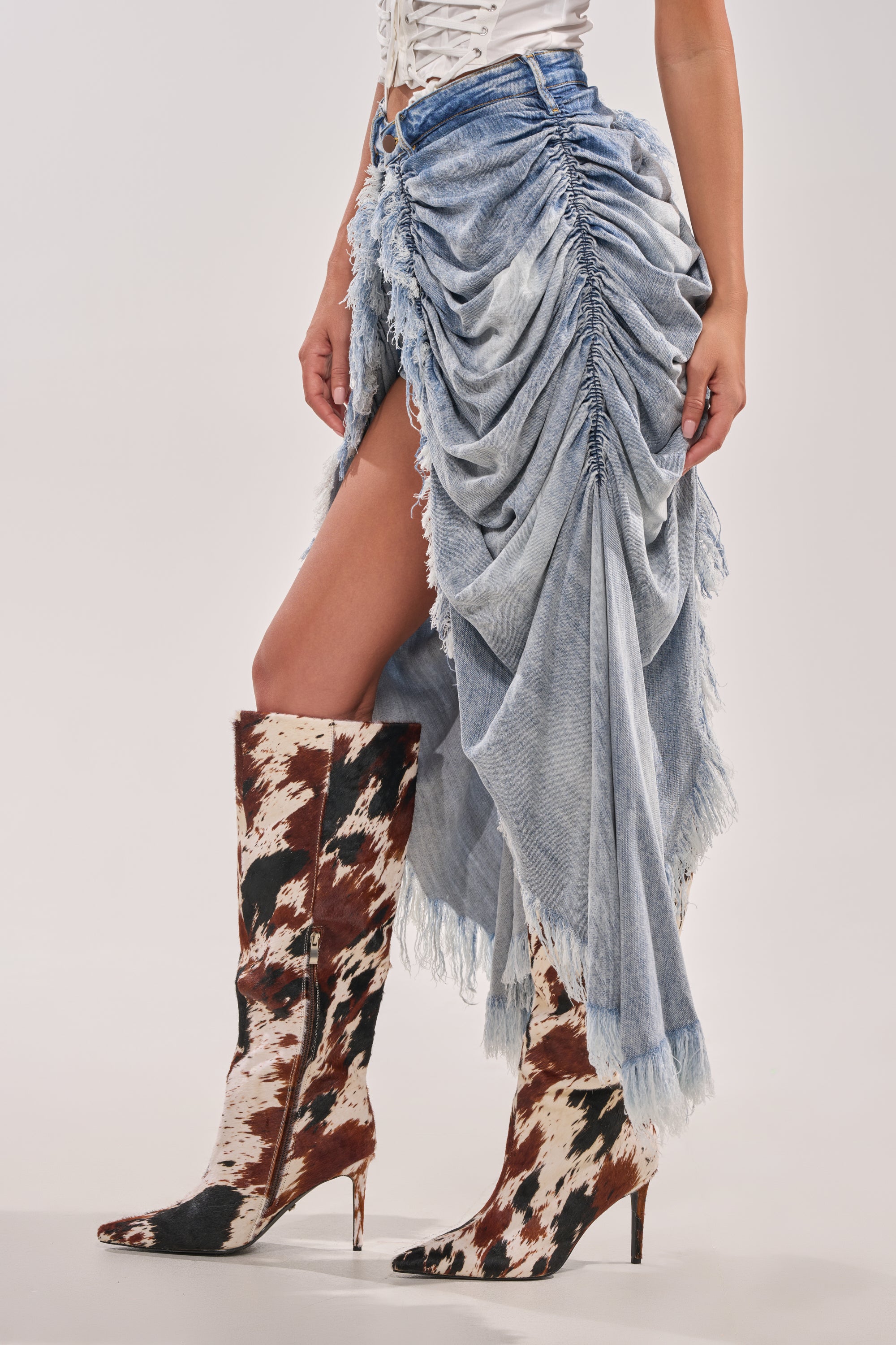 Someone wearing the ALL OVER THE PLACE DENIM SHORT—with ruched details, high-rise waist, distressed finish, and a high slit—paired with knee-high, pointed-toe boots in a brown and white cow print. 3