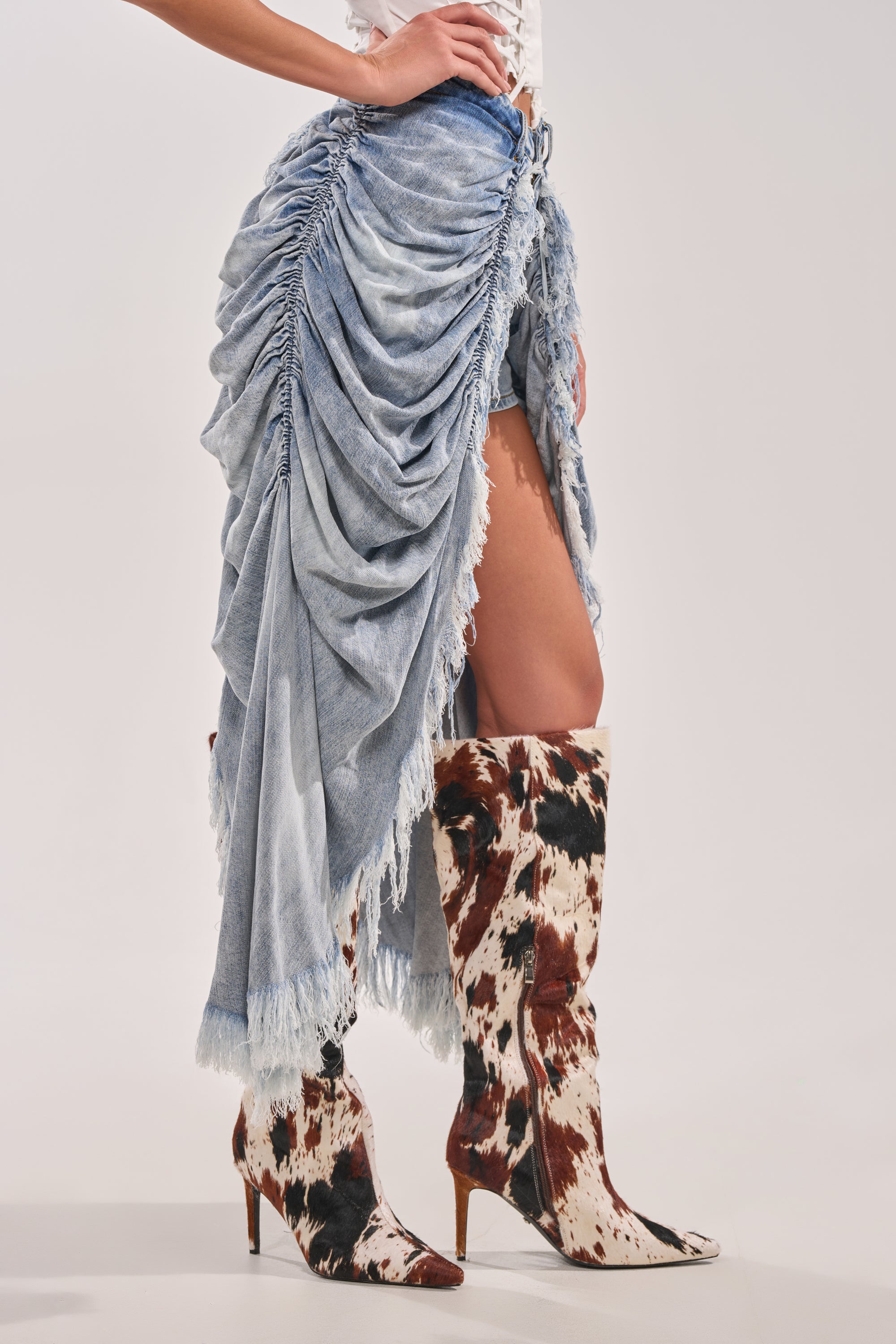 A person stands in cow-print heeled boots and a ruched denim skirt with a high slit, revealing the ALL OVER THE PLACE DENIM SHORT underneath. Their hand rests on their hip against a plain background. 4