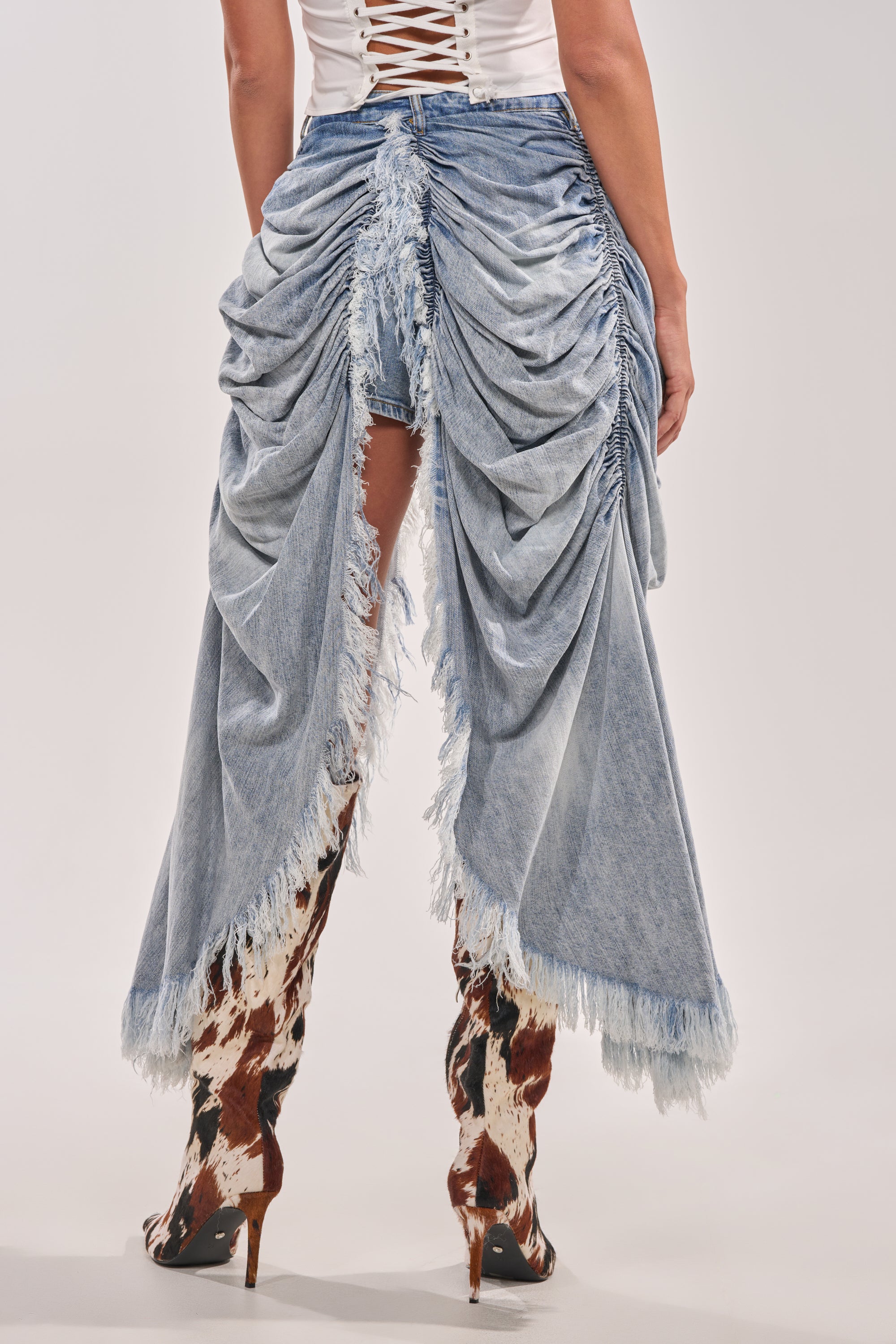 Wearing the ALL OVER THE PLACE DENIM SHORT—high-rise, draped and ruched with frayed edges and distressed details—styled with a white lace-up top and brown-and-white cow print heeled boots. Photo is from the back, showcasing the denim’s design. 5
