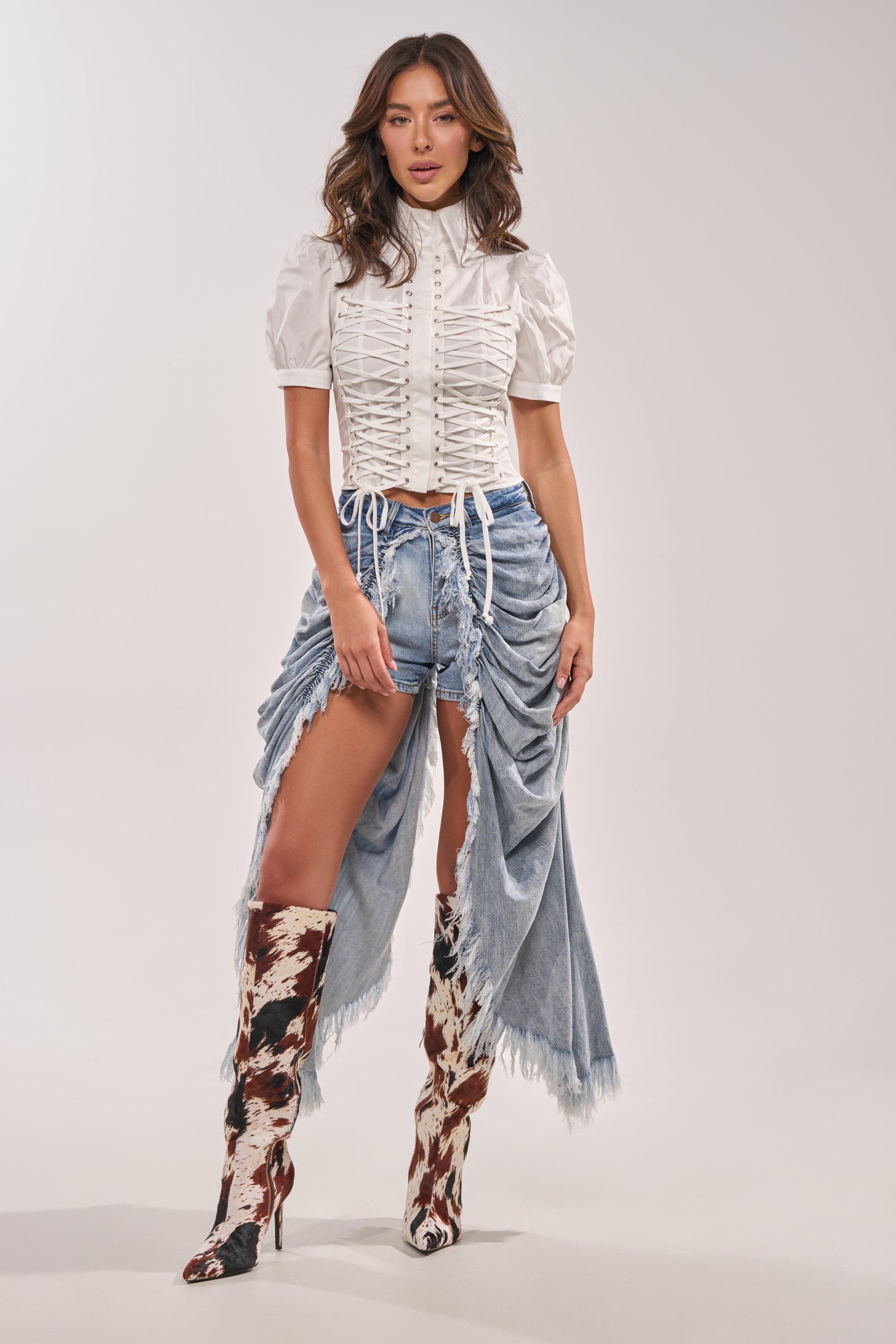 A woman poses in the ALL OVER THE PLACE DENIM SHORT—high-rise, distressed shorts with long ruched denim panels—styled with a white ruched blouse and knee-high brown and white cow-print boots against a plain background. 6