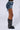 A person's lower half is displayed wearing the DONT DO IT DENIM SHORT, featuring heavy distressing and a high-rise waist, paired with black knee-high boots that are embellished with silver chains and studs. The simple white background serves to accentuate the daring fashion statement.