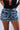 A person wears the "DONT DO IT DENIM SHORT" featuring distressed detailing, showcasing multiple horizontal rips that reveal frayed fabric beneath. They pair the shorts with a black top adorned with metal pins and a red cross design. Their hands rest casually on their hips.