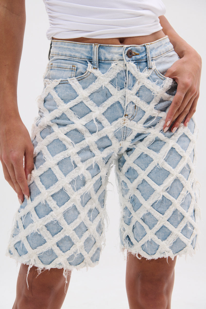 A person wears the DISTRESSED AND STRESSED DENIM BERMUDA SHORT—high-rise, light blue, with a white textured lattice overlay and frayed edges—paired with a white shirt. The photo shows the shorts from waist to just above the knees.
