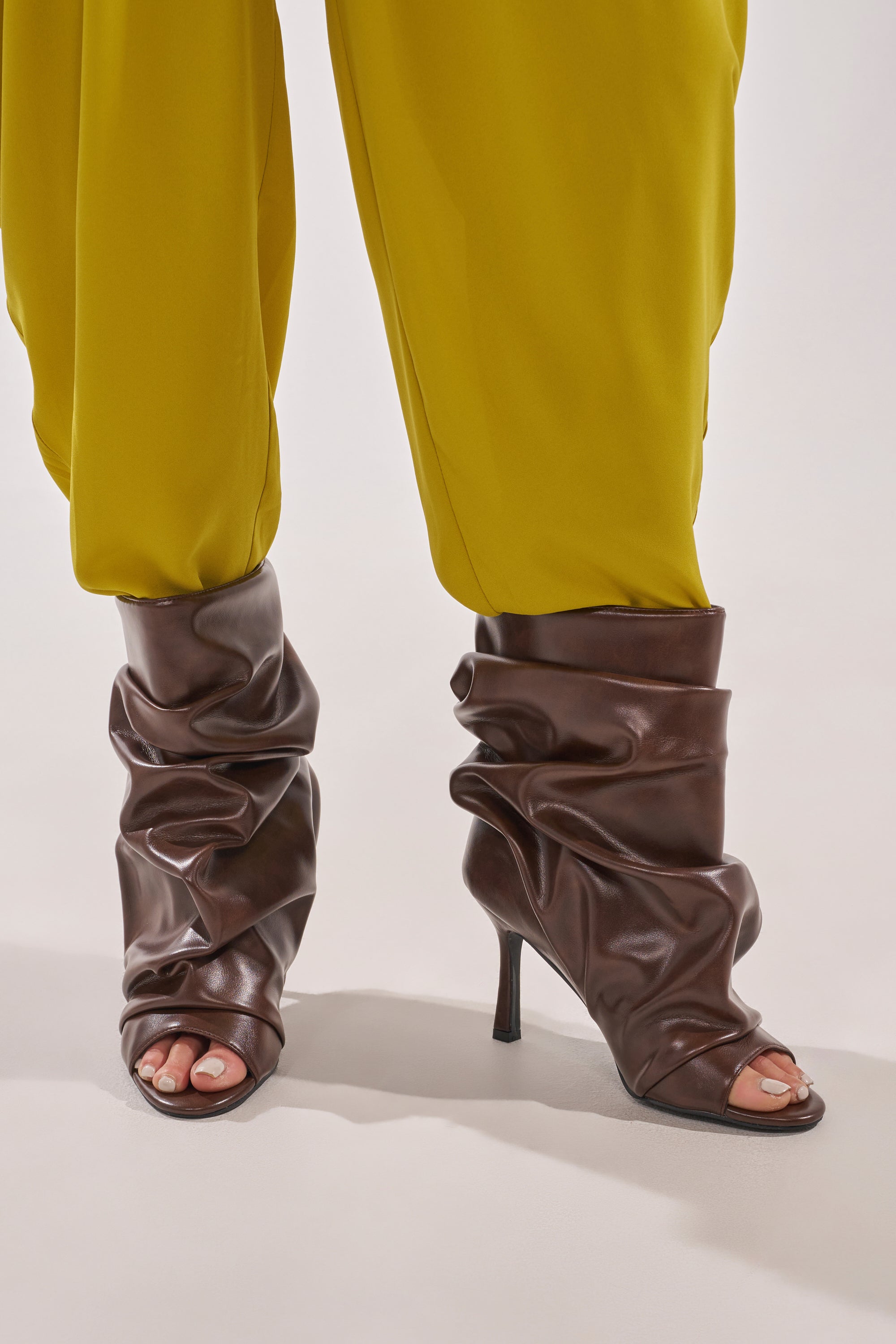A person wears loose yellow pants tucked into AZALEA WANG APEX BROWN SLOUCHY OPEN TOE BOOTIE stiletto booties, standing on a light-colored surface. 0