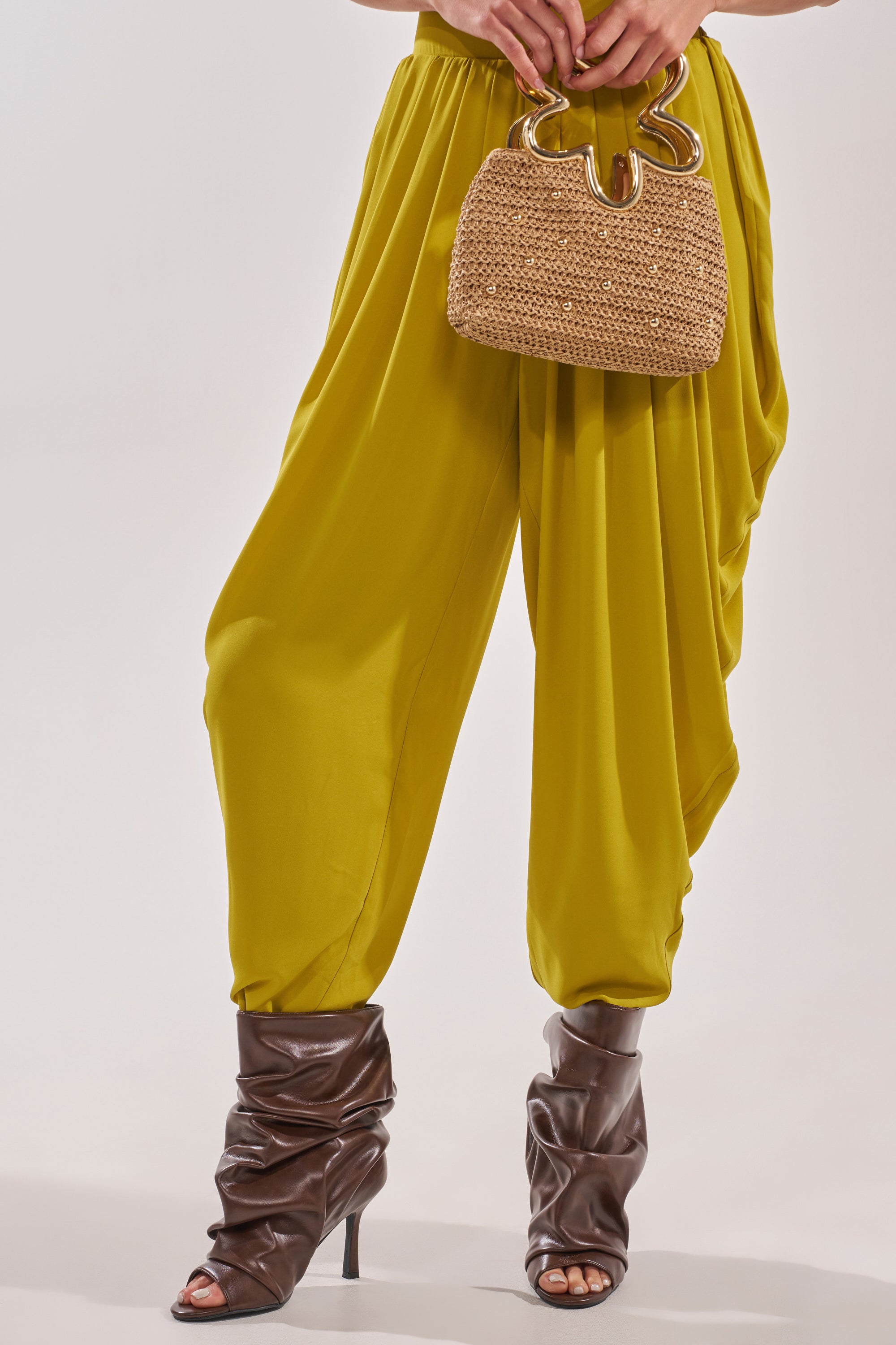 A person wearing loose yellow harem pants, the AZALEA WANG APEX BROWN SLOUCHY OPEN TOE BOOTIE, and holding a tan woven handbag with gold handles, stands against a plain light background. 1