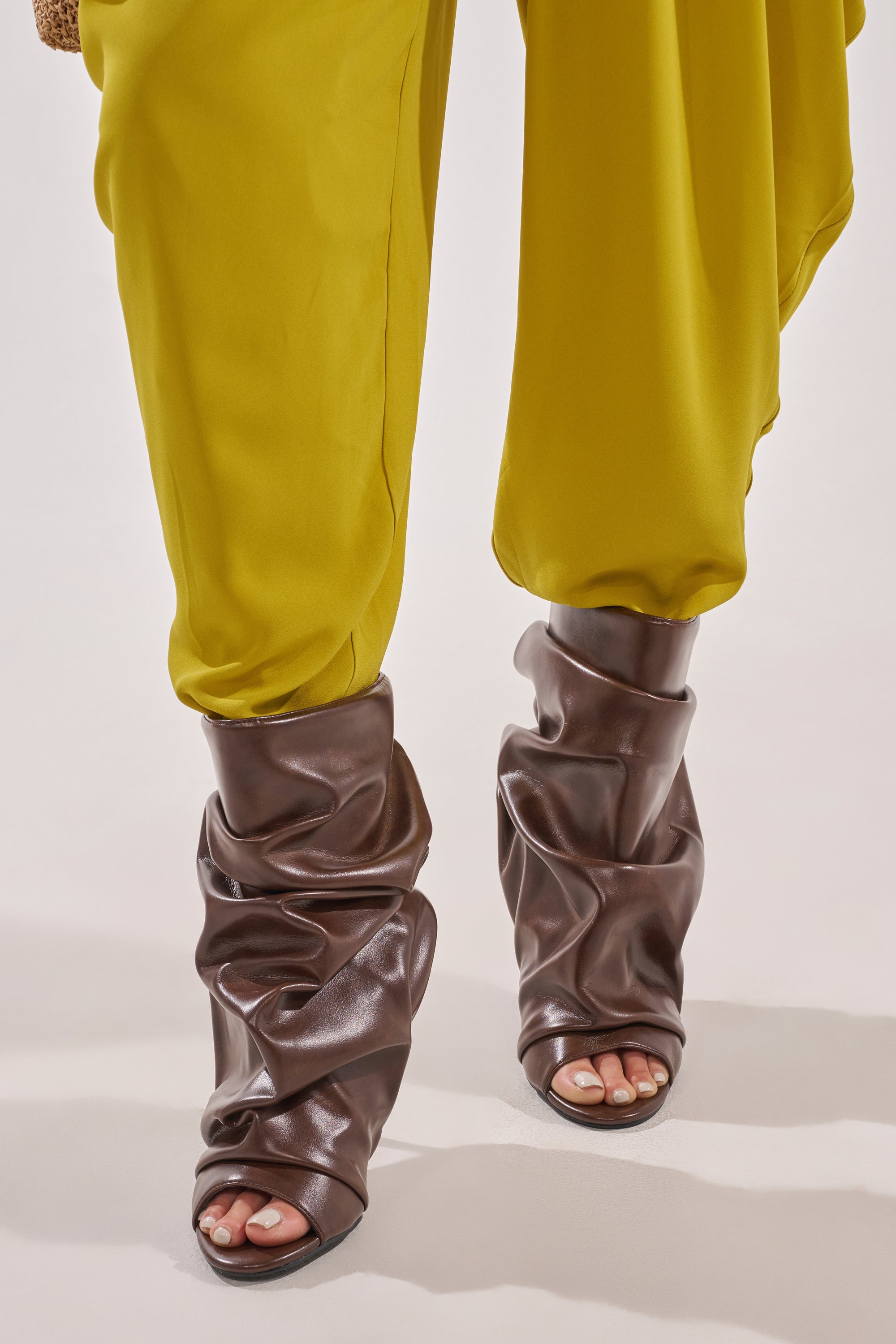 A person wears loose, shiny yellow pants tucked into AZALEA WANG APEX BROWN SLOUCHY OPEN TOE BOOTIE high heels, standing on a light-colored surface. Only the lower legs and feet are visible. 2