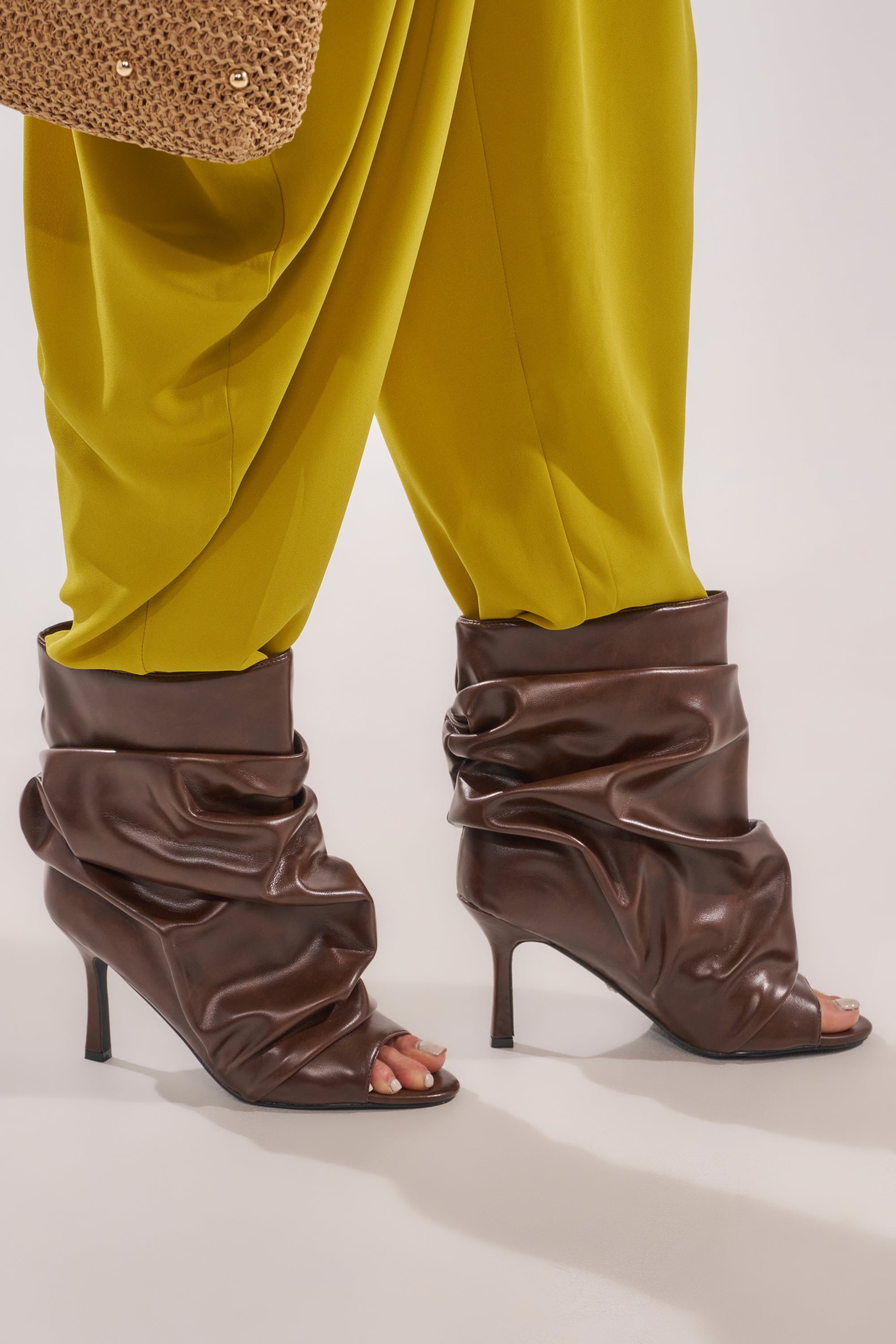 Someone wears loose, bright yellow pants tucked into AZALEA WANG APEX BROWN SLOUCHY OPEN TOE BOOTIE heels. Part of a woven tan handbag appears at the top left. 3