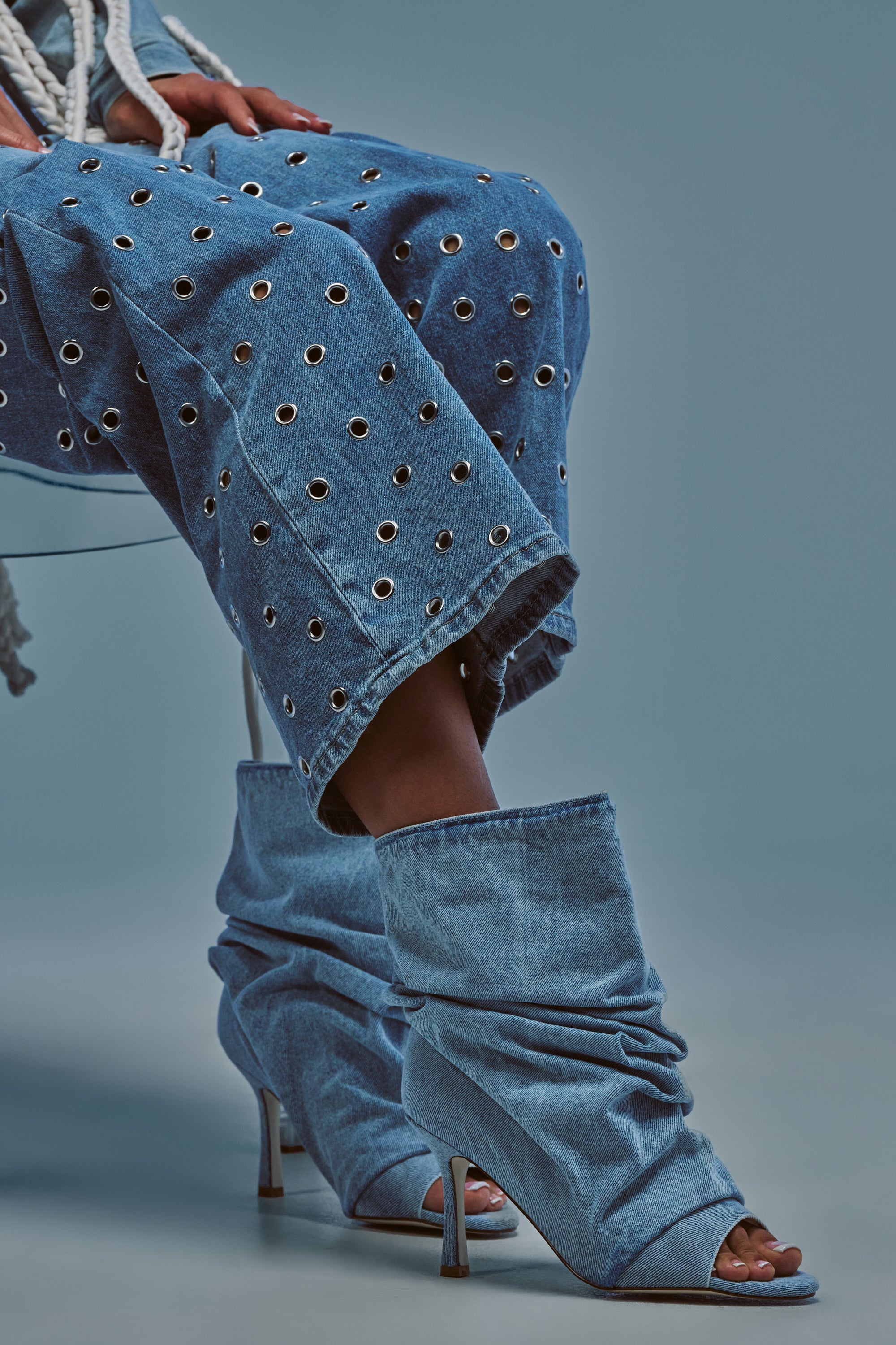 Wearing wide-leg denim pants with metal eyelets and the AZALEA WANG APEX DENIM RUCHED STILETTO BOOTIE, a person showcases their fashion-forward edge with statement footwear against a plain blue background.