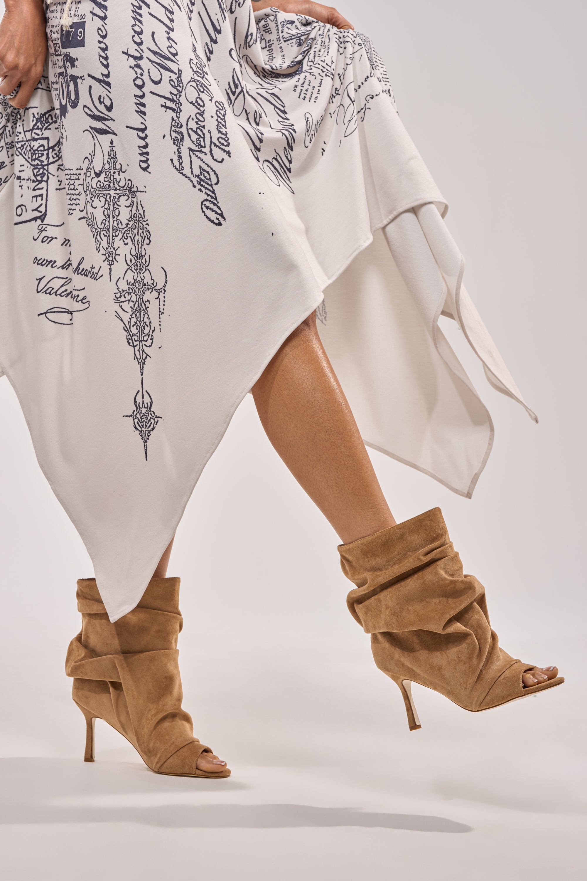 A person stands on tiptoe, one leg lifted, wearing a white skirt with black script designs and showcasing the AZALEA WANG APEX LIGHT BROWN FAUX SUEDE OPEN TOE BOOTIE against a plain background. 2